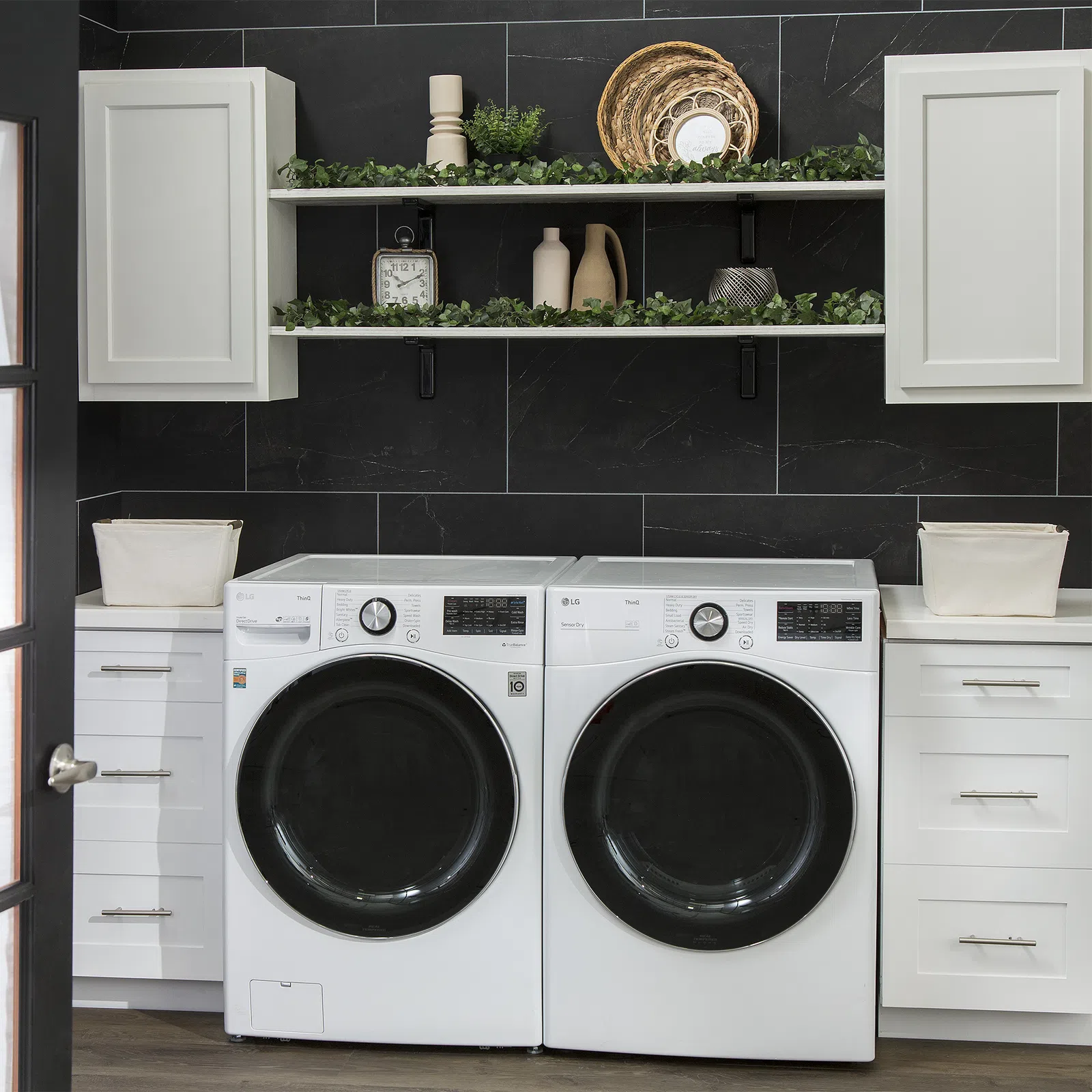 Modern laundry room with white front-load washer and dryer, black tiled wall, white cabinets, and decorative shelves with plants.