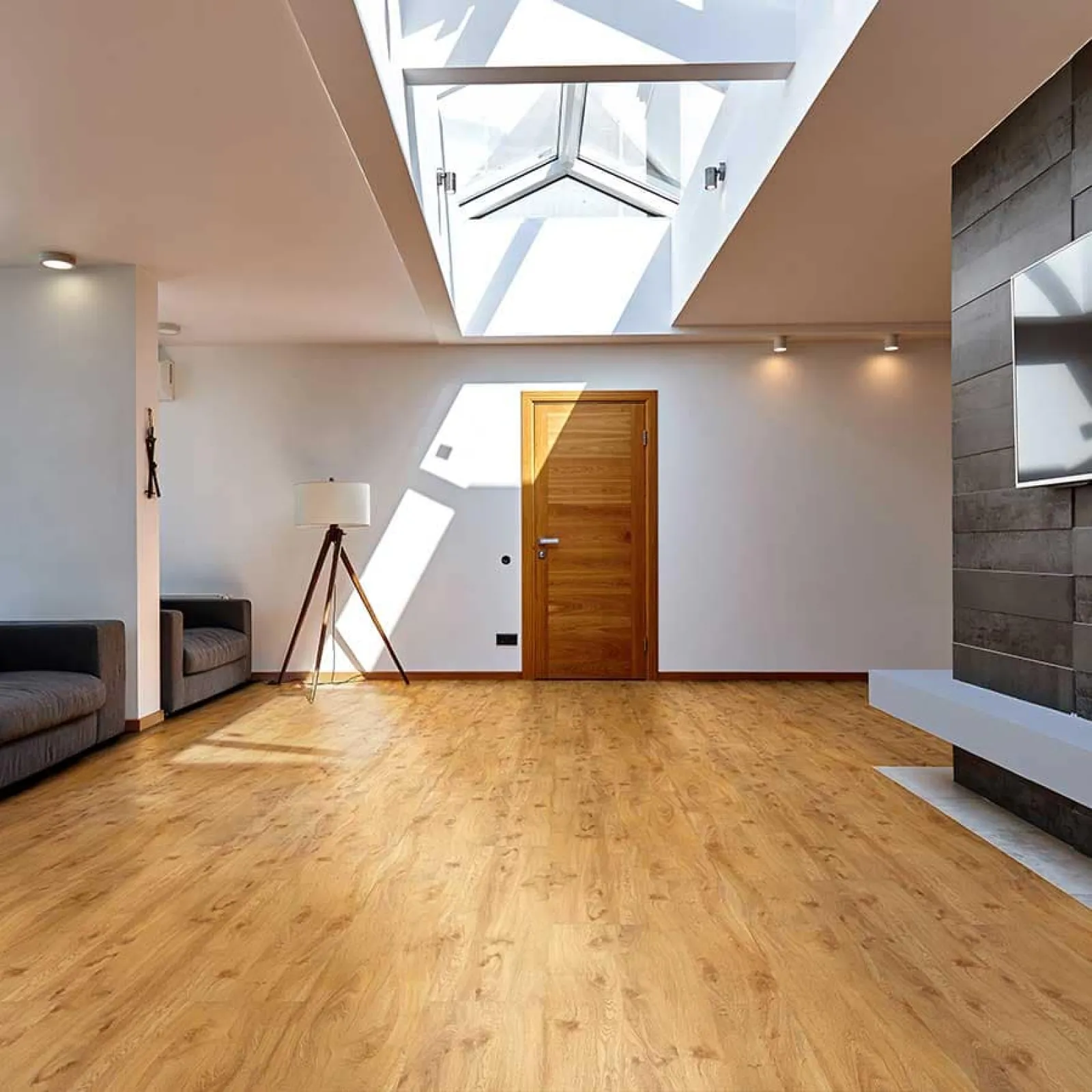 Bright modern living room with wooden flooring, skylight, wooden door, gray sofas, and a tripod floor lamp.
