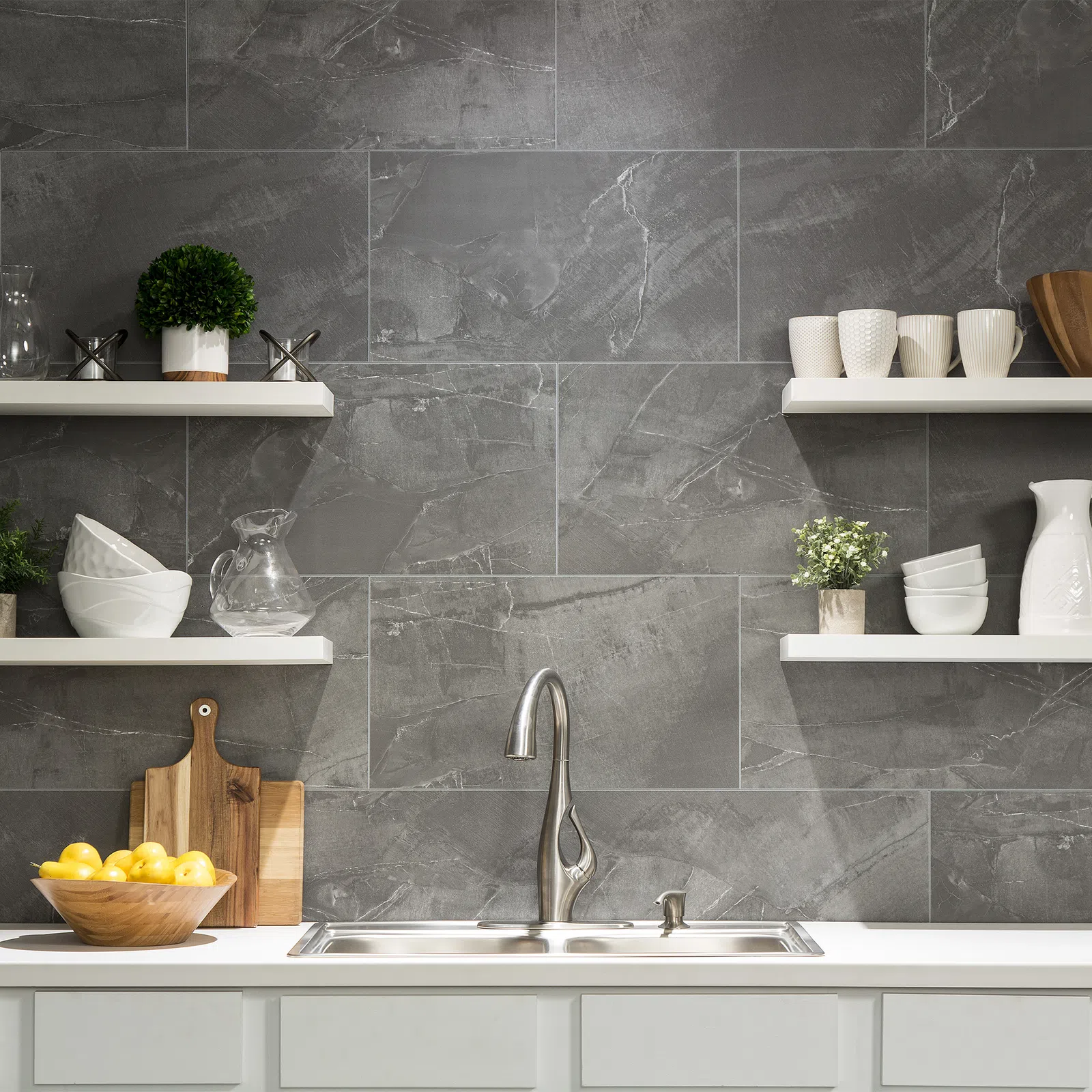 Modern kitchen sink with stainless steel faucet, gray stone backsplash, white shelves with dishes and plants.