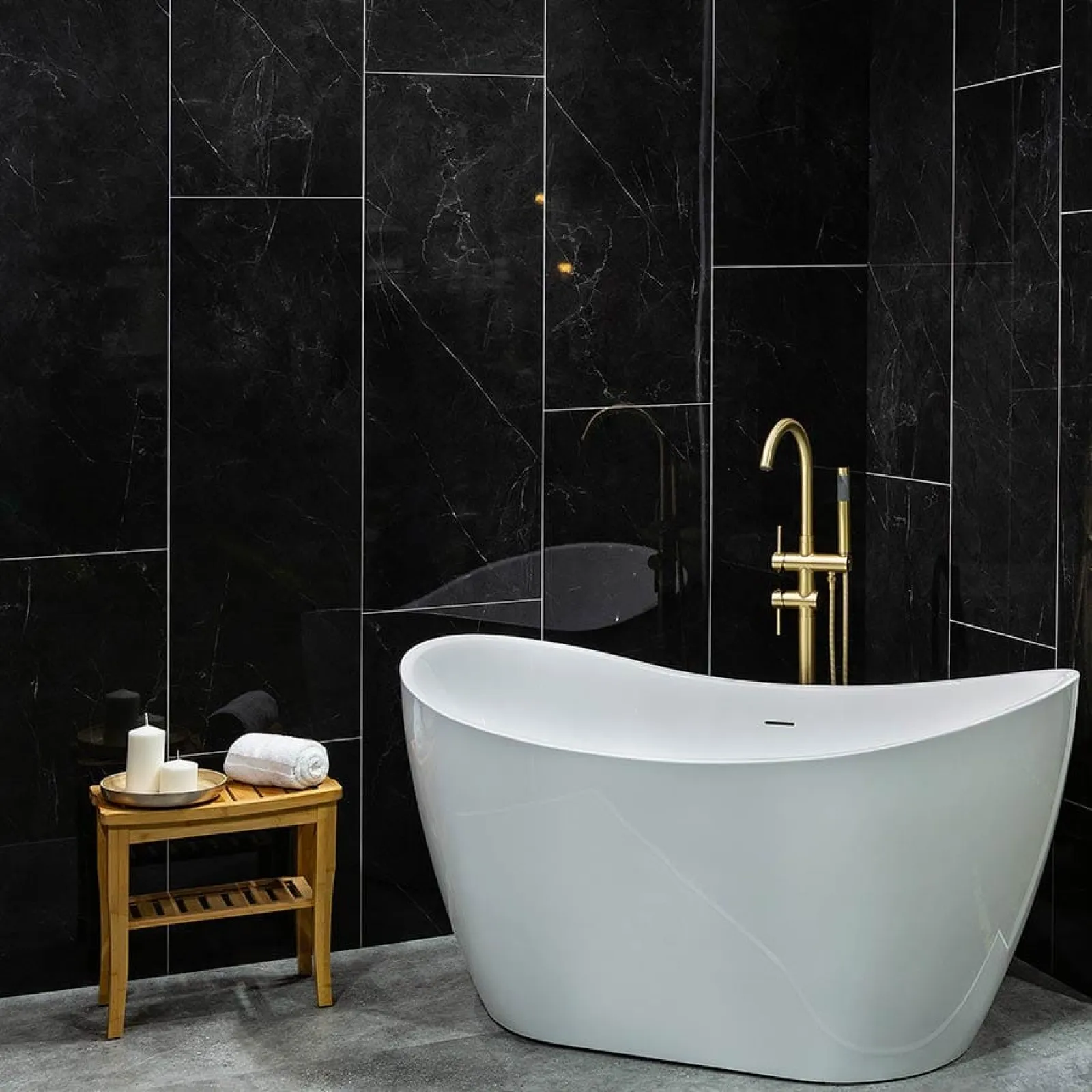 Modern white freestanding bathtub with gold faucet against sleek black marble wall with wooden side table.