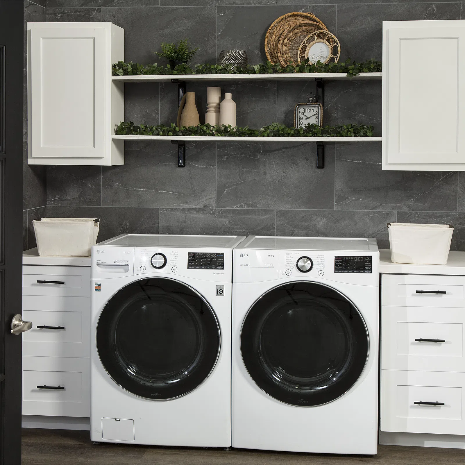 Modern laundry room with white front-load washer and dryer, white cabinets, shelves with decor, and gray tiled wall.