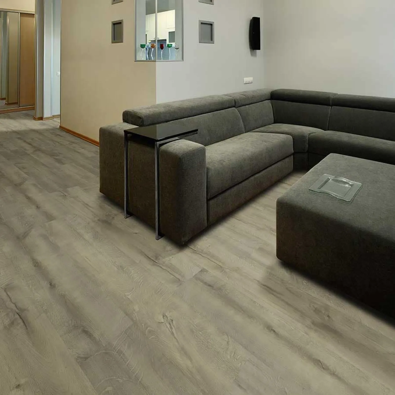 Modern living room with gray sectional sofa, dark ottoman, and light wood laminate flooring under soft lighting.