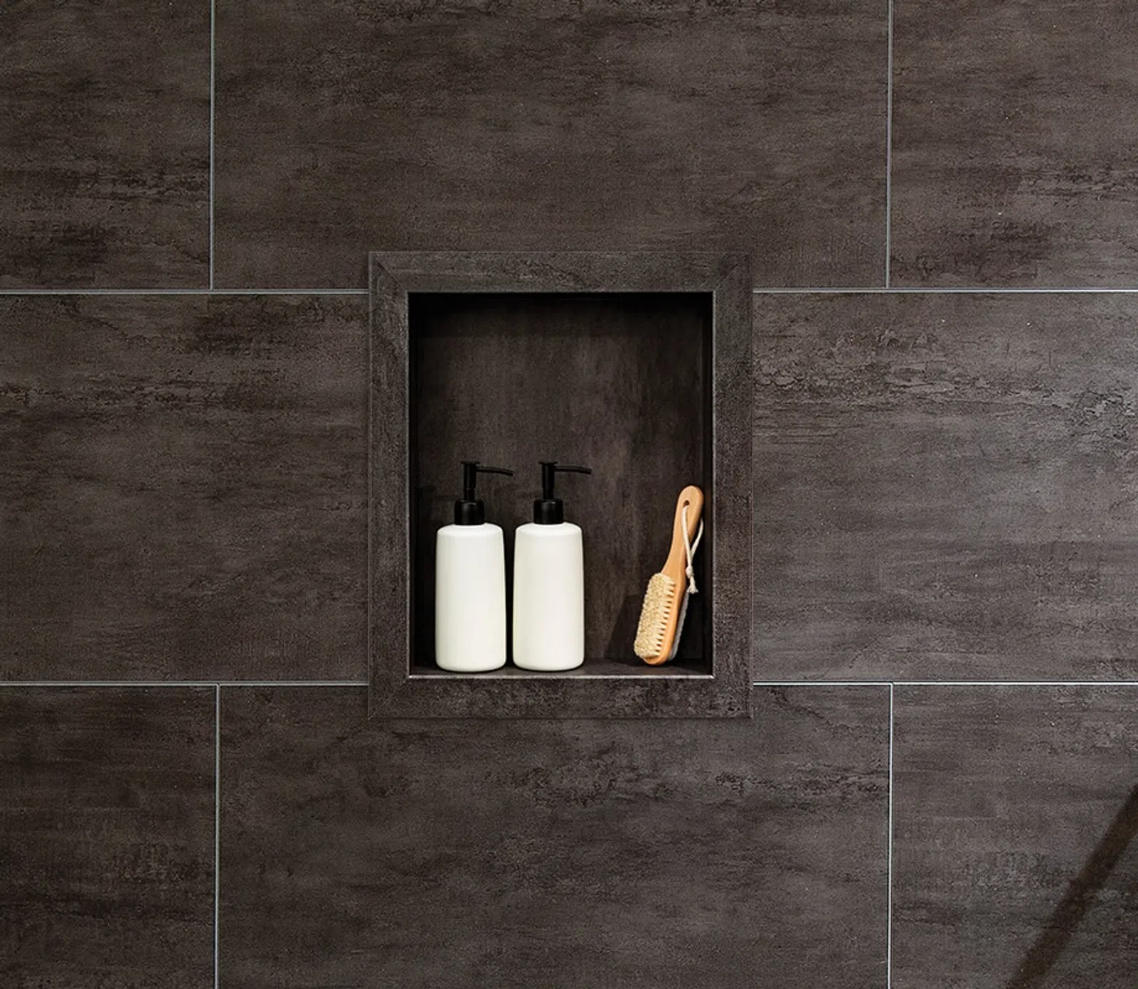 Dark tiled shower niche with two white pump bottles and a wooden bath brush neatly arranged inside