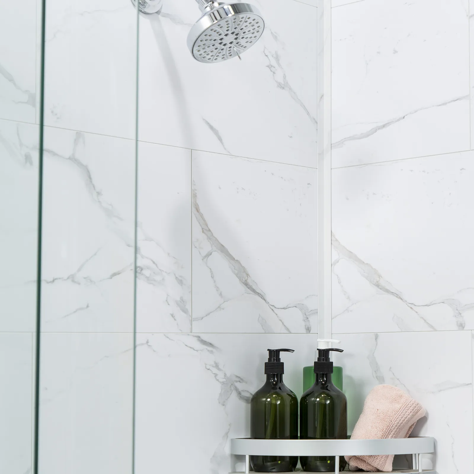 Modern showerhead with glass shower screen, marble wall tiles, green soap dispensers, and a folded towel on a rack.