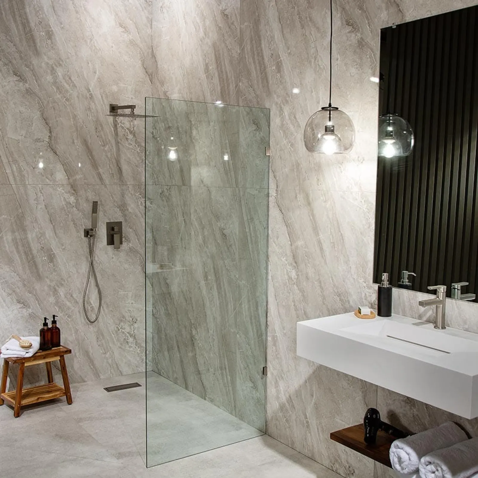 Modern bathroom with glass shower enclosure, marble walls, floating sink, pendant light, and wooden stools.