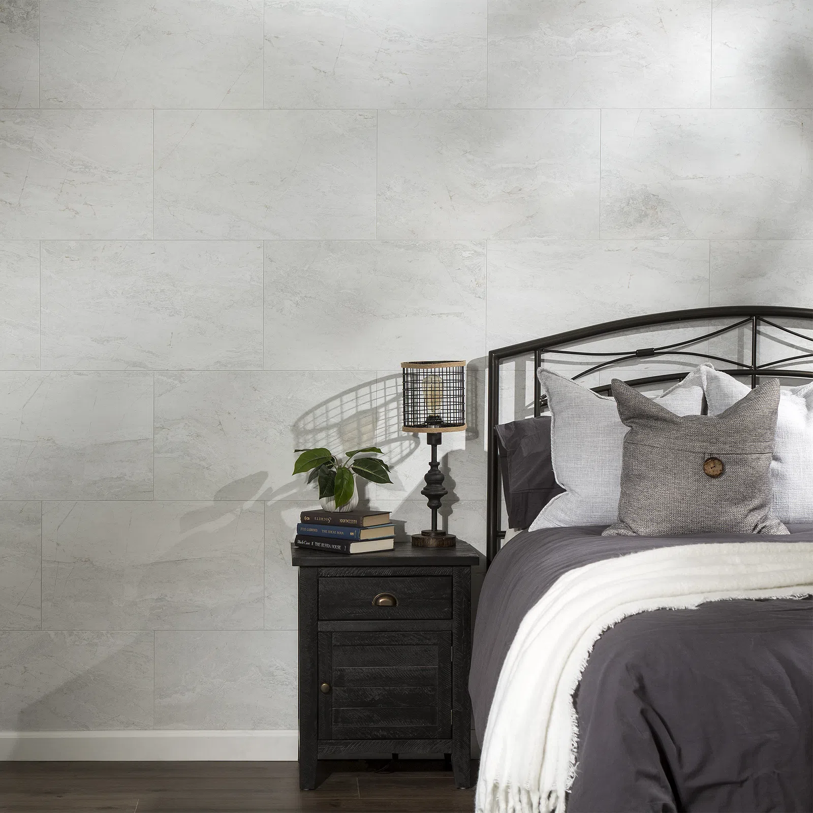 Cozy bedroom corner with dark metal bed, gray bedding, black nightstand, lamp, books, and plant against light stone wall