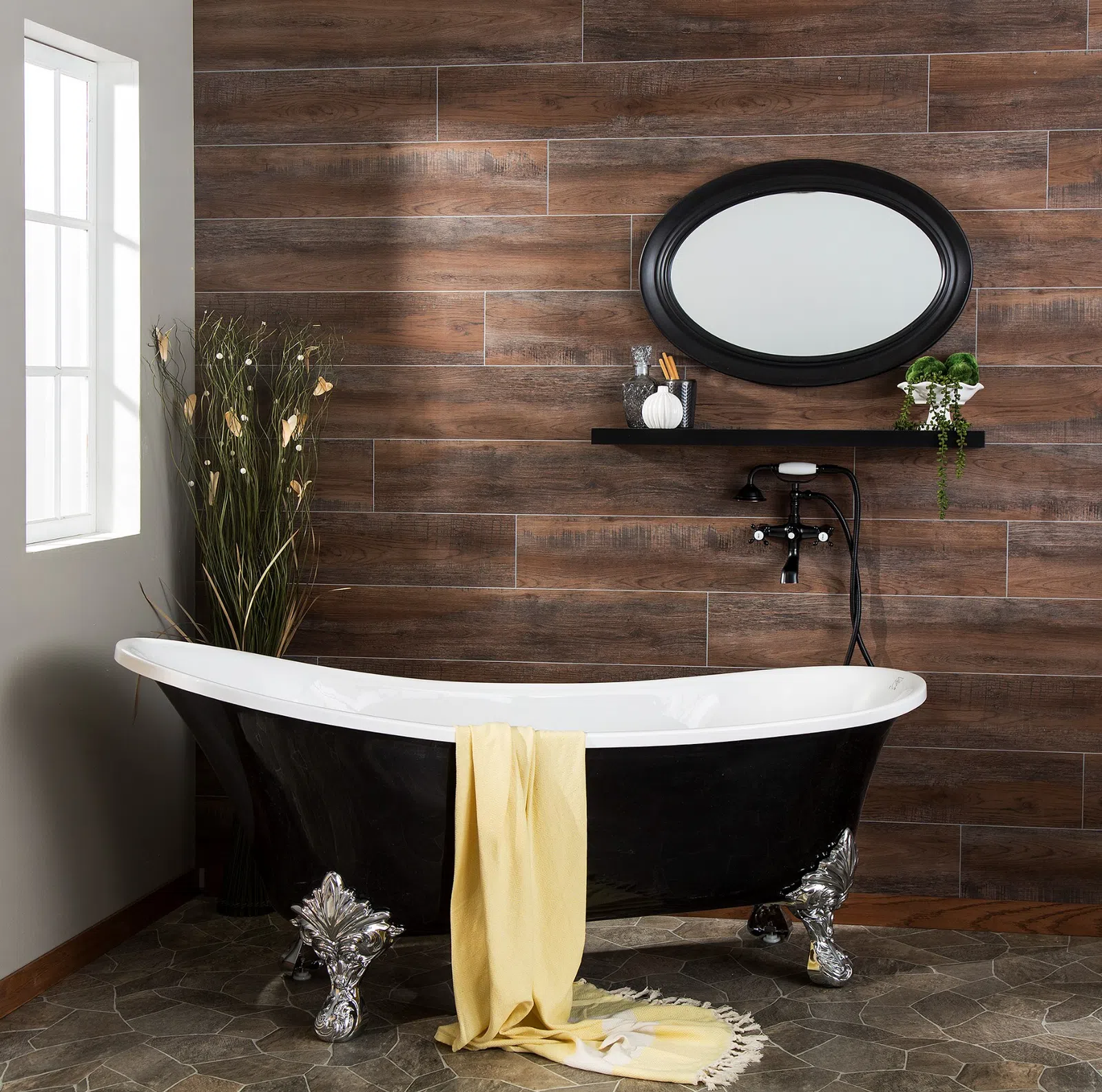 Black clawfoot bathtub with yellow towel in a rustic bathroom featuring wood wall tiles and oval mirror