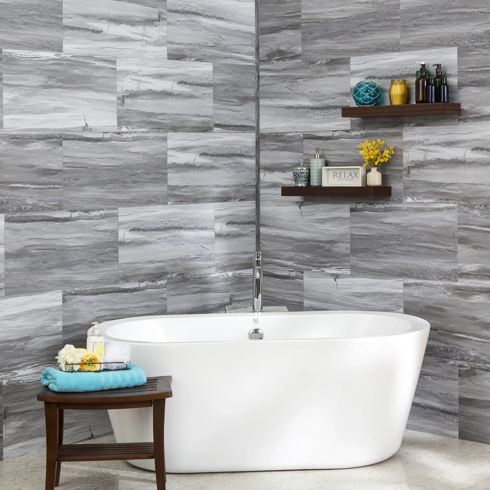 Modern white freestanding bathtub with gray stone tile walls and decorative shelves in bathroom corner.