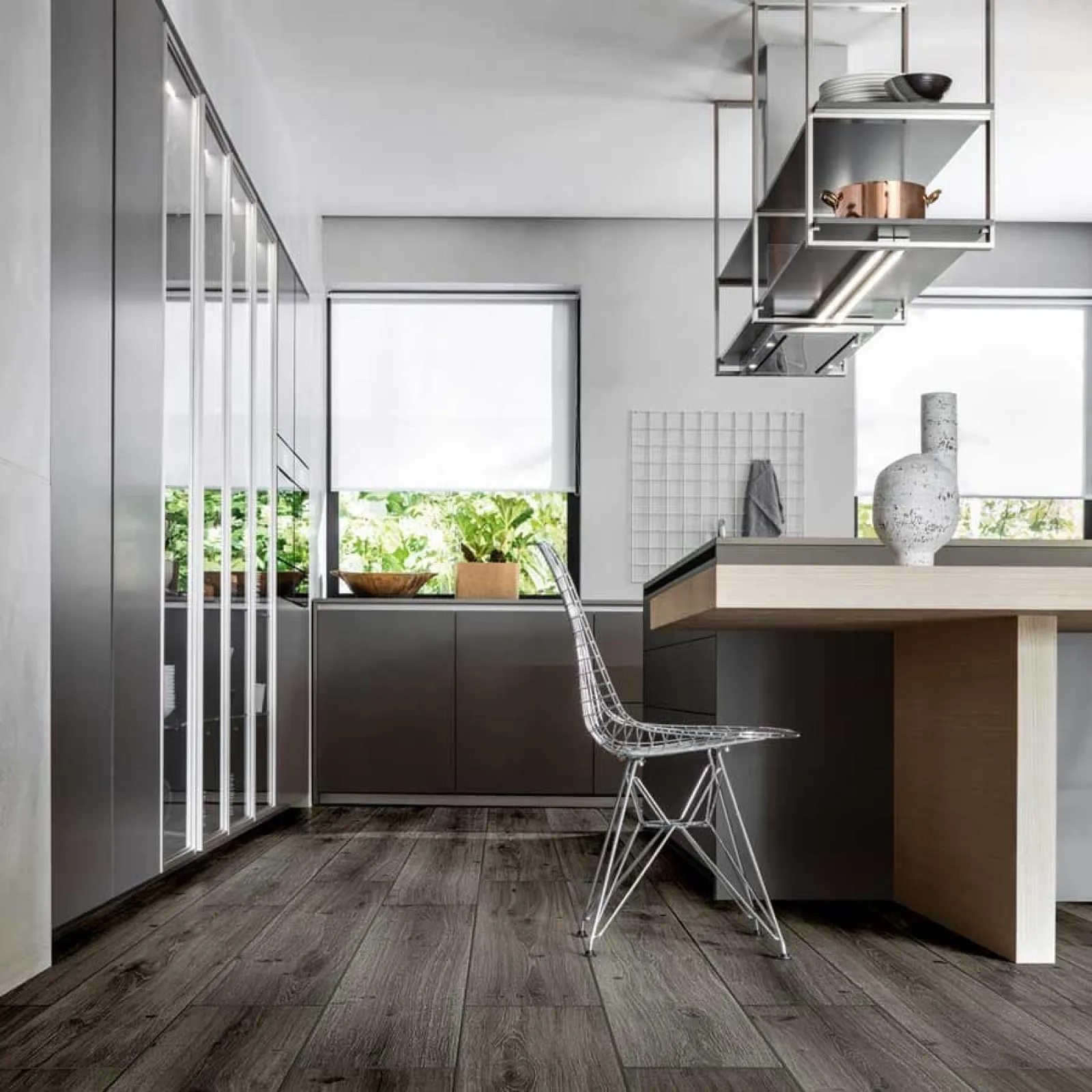 Modern kitchen with sleek gray cabinets, wooden flooring, metal wire chair, and minimalist decor under natural light.