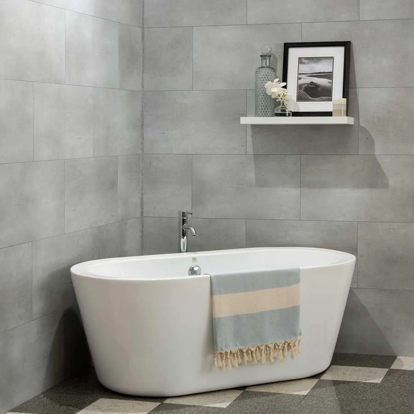 Modern white freestanding bathtub with a striped towel in a gray tiled bathroom with a floating shelf above.