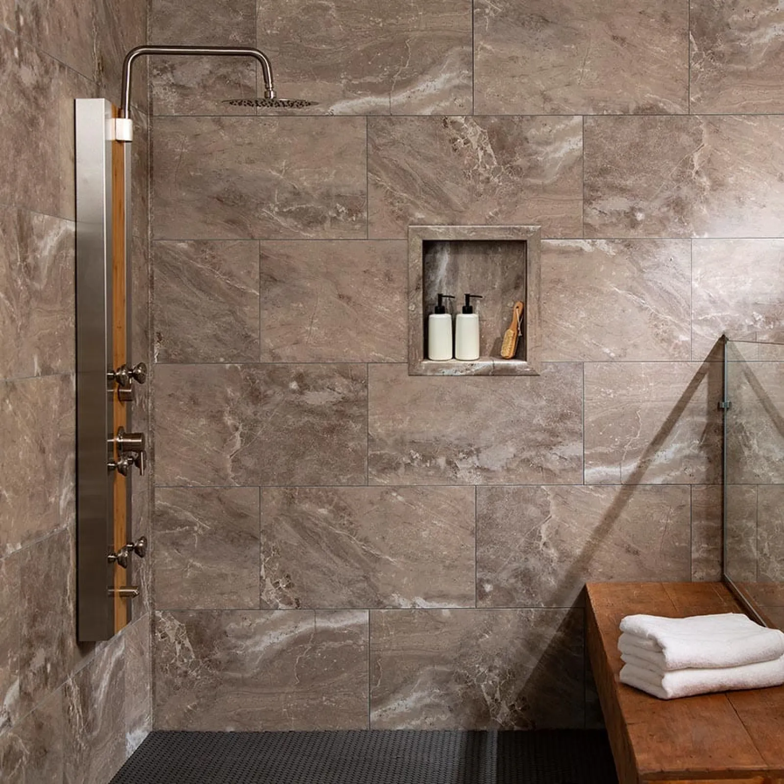 Modern shower area with large stone tiles, rain showerhead, built-in shelf with toiletries, and wooden bench with towels.