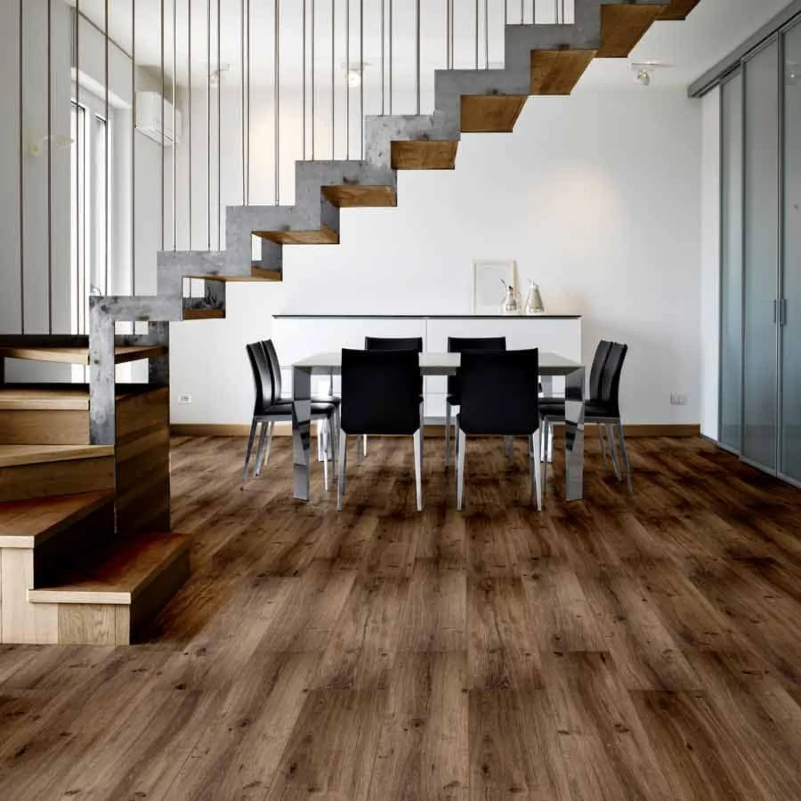 Modern dining room with wooden floors, black chairs, glass table, and industrial staircase with metal railings.