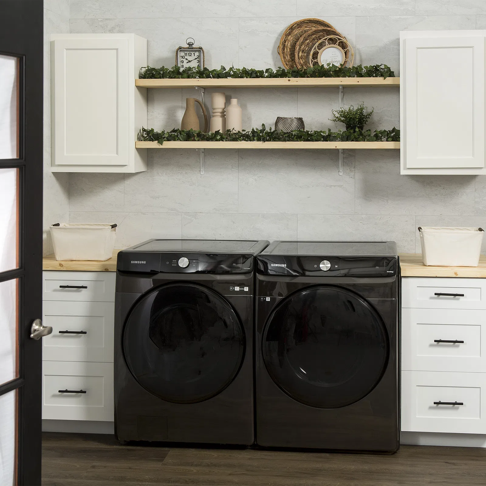 Modern laundry room with black Samsung washer and dryer, white cabinets, wooden shelves, and decorative greenery.