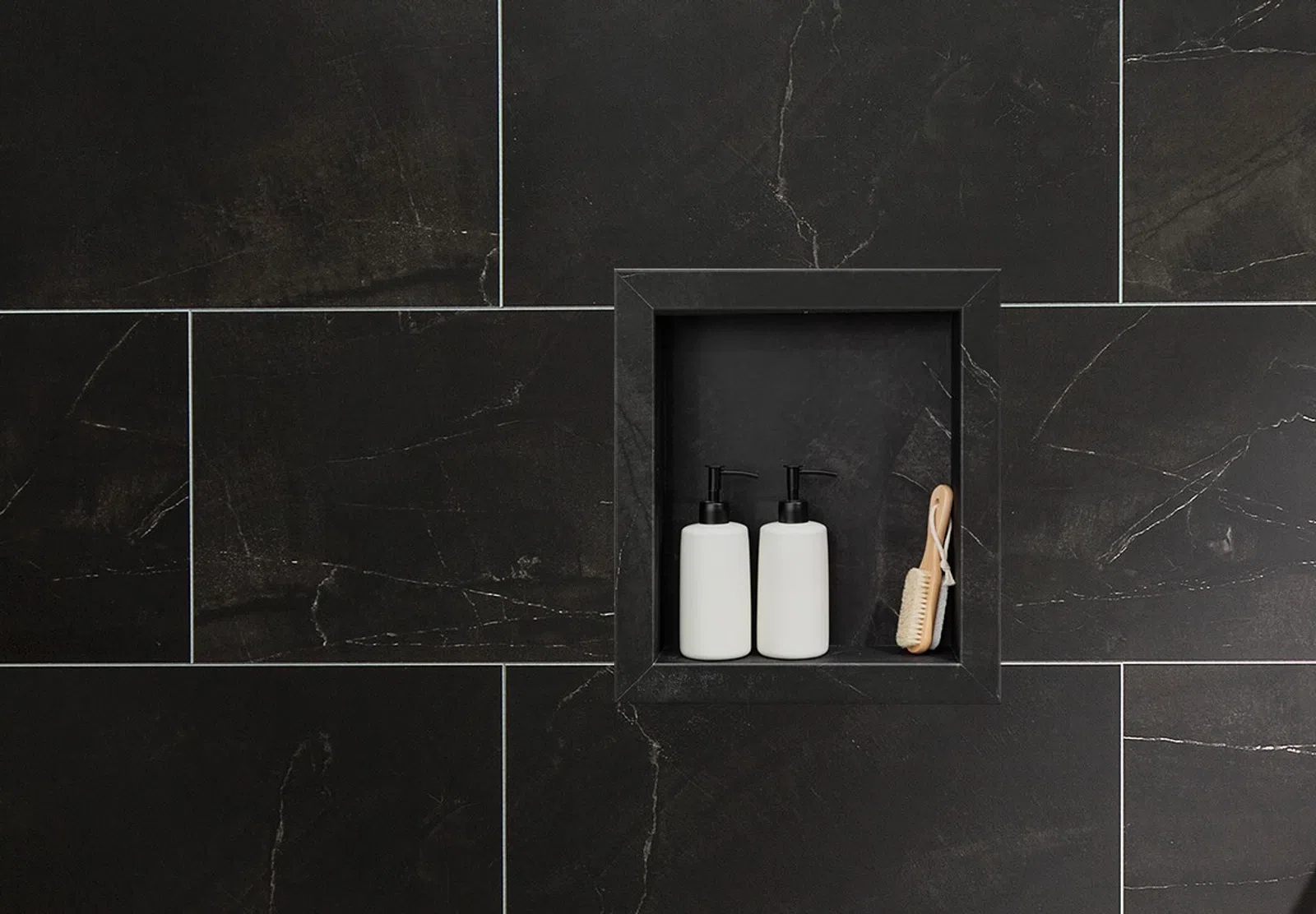 Two white pump bottles and a wooden brush in a black tiled shower niche with dark grout lines.