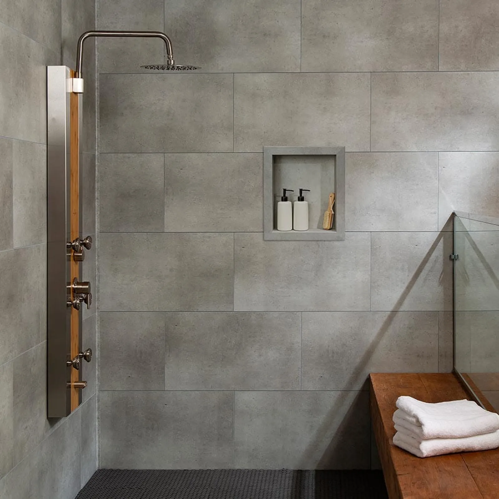 Modern shower area with gray tiled walls, built-in soap niche, wooden bench, and folded towels on bench.