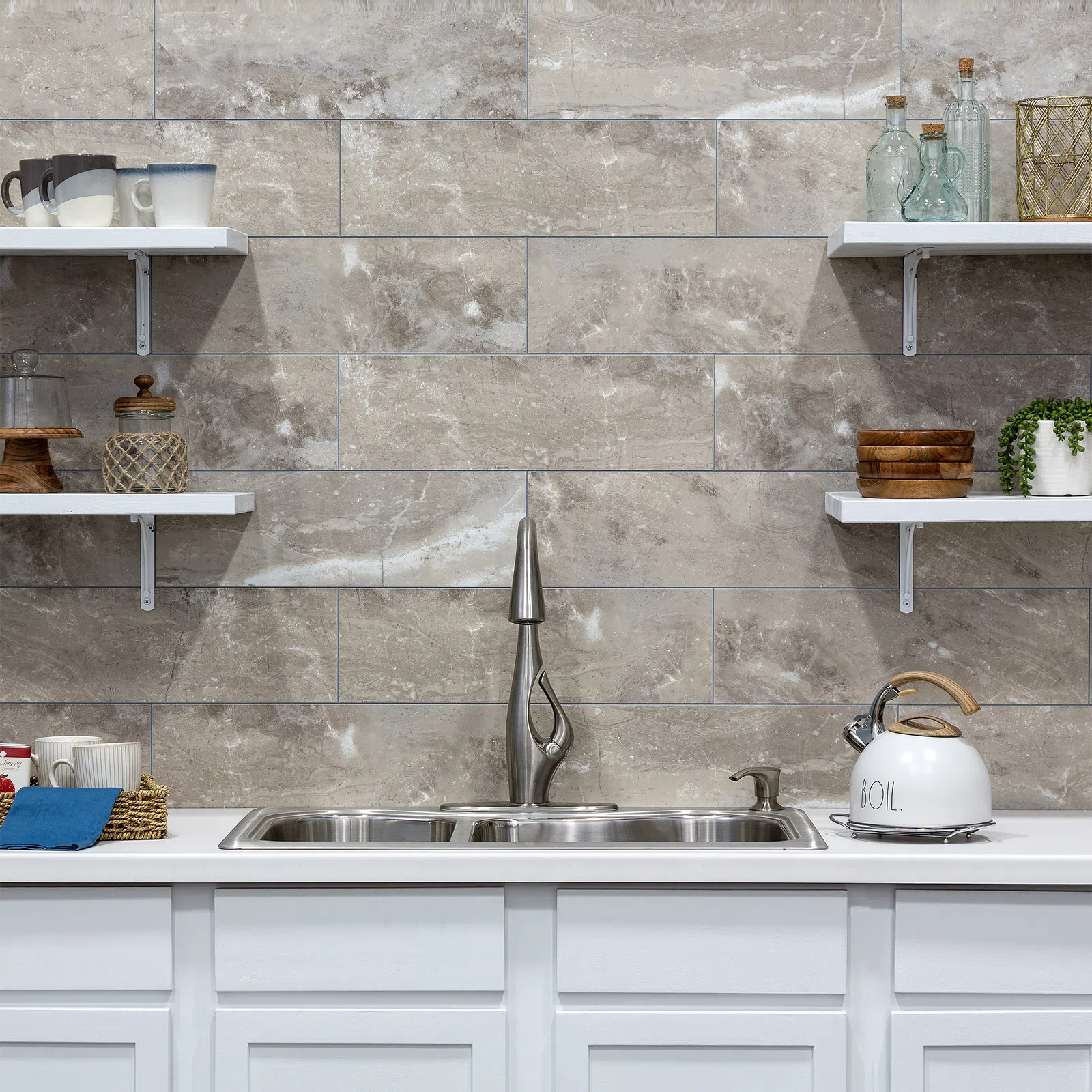 Modern kitchen sink with stainless steel faucet, white countertop, gray tile backsplash, and open shelves with decor.