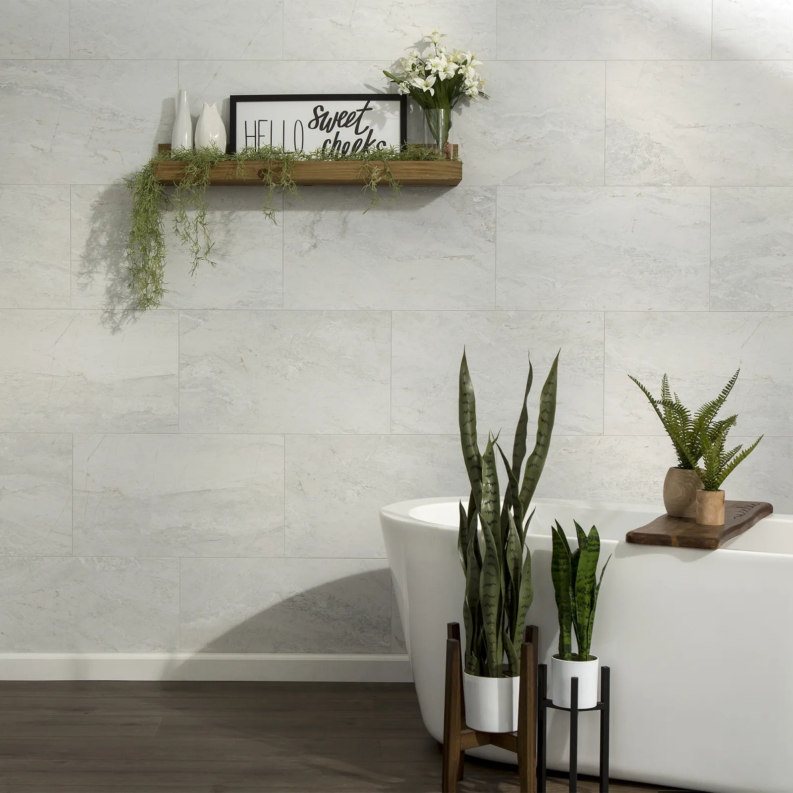 Modern bathroom with freestanding white tub, green plants, floating wooden shelf, and light gray tiled wall.