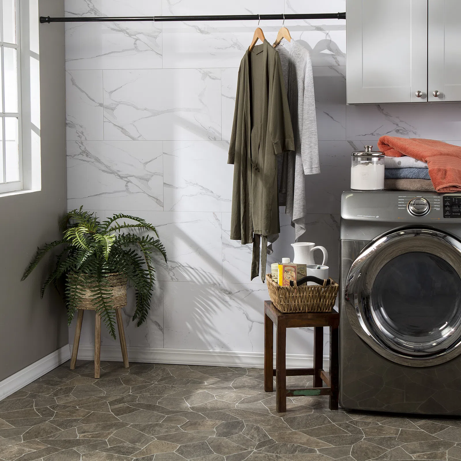 Modern laundry room with front-load washer, hanging robes, wicker plant stand, and folded clothes on cabinet