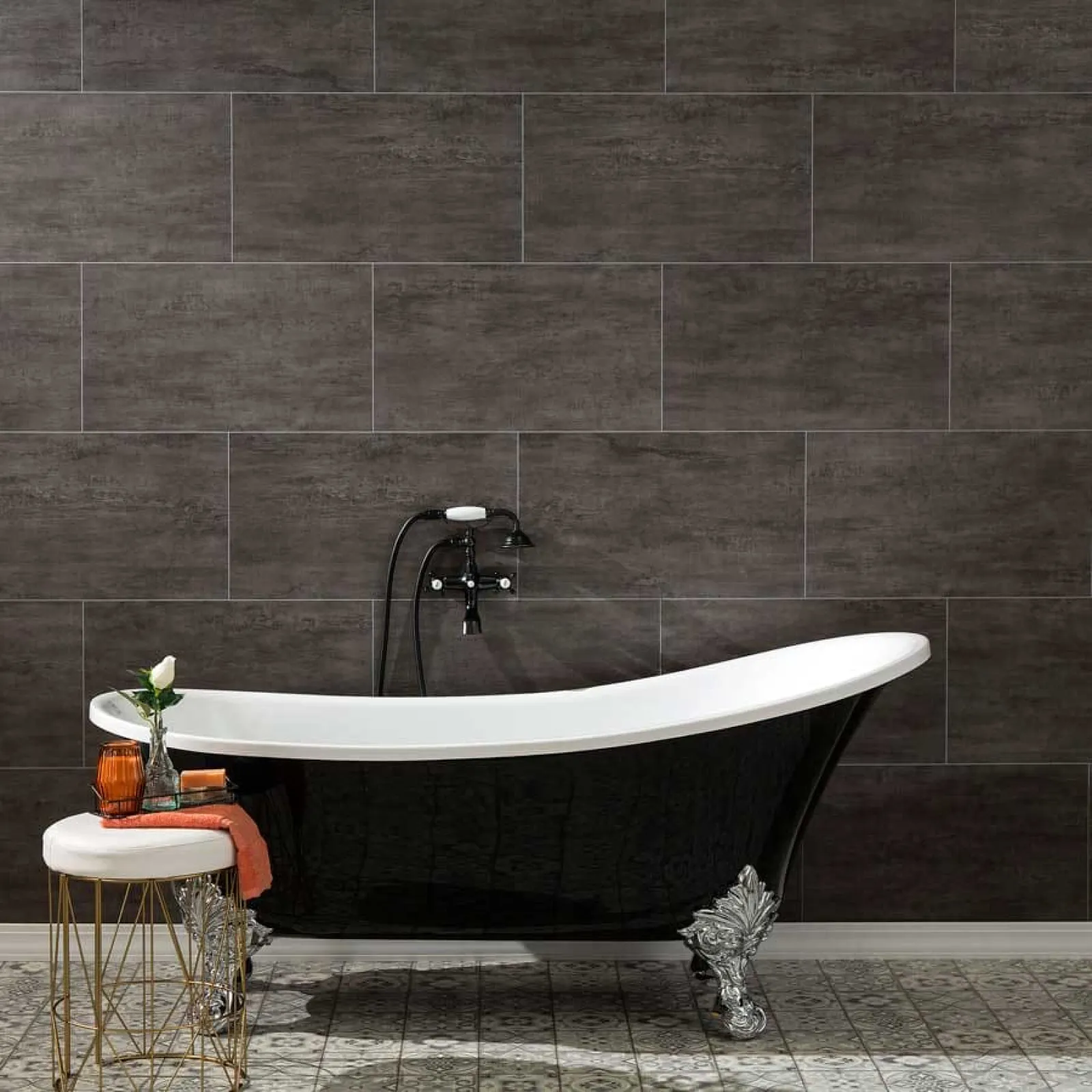 Black clawfoot bathtub with silver feet against dark tile wall and decorative stool with orange towel and vase