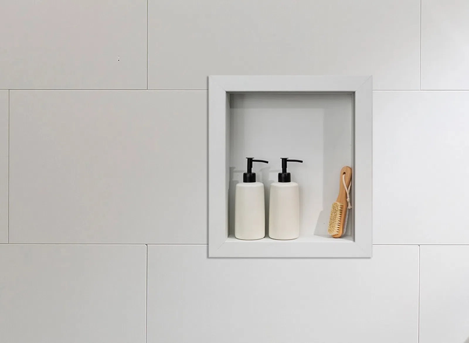 White tiled bathroom wall with recessed shelf holding two white pump bottles and a wooden brush.