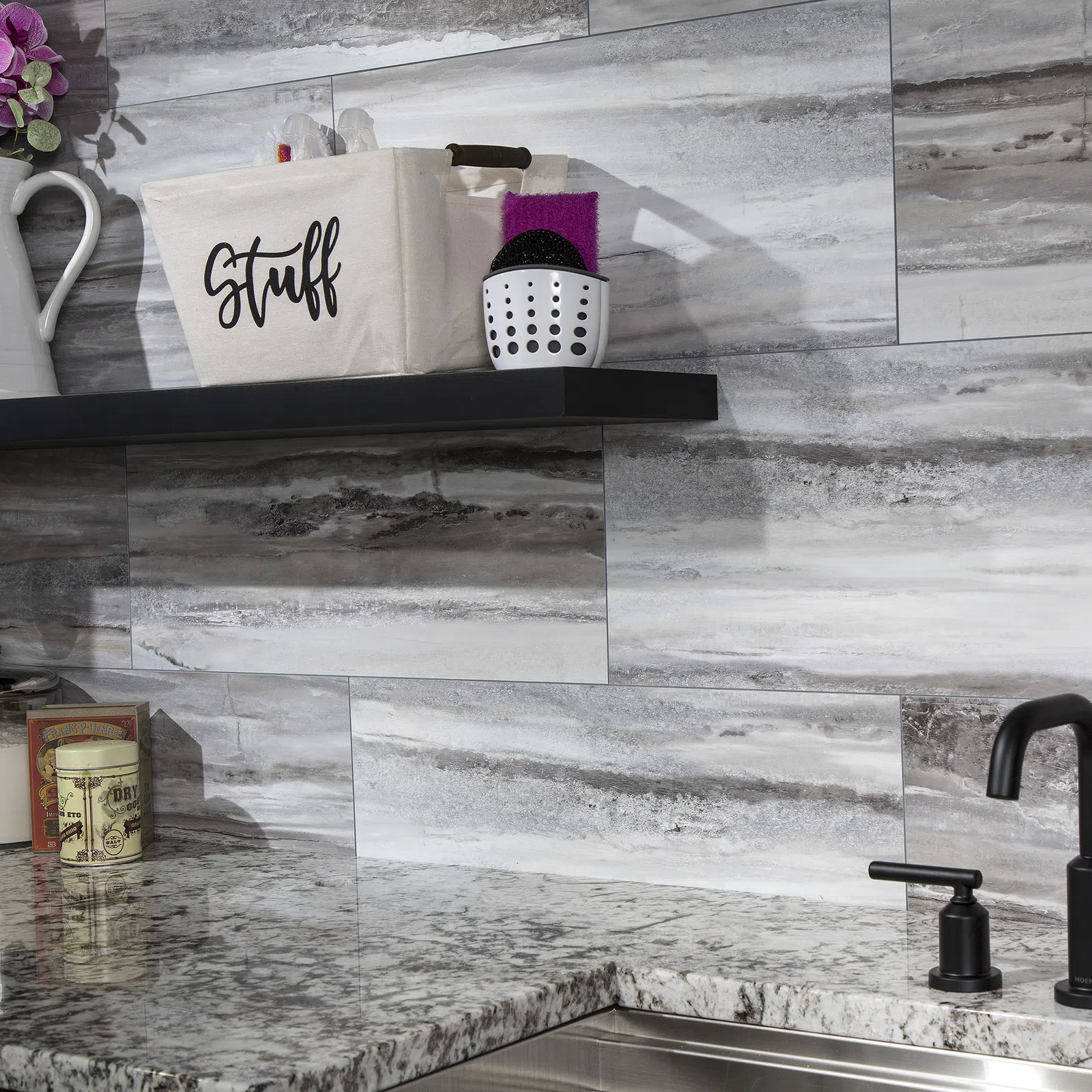 Modern kitchen corner with gray stone backsplash, granite countertop, black faucet, and storage basket labeled Stuff