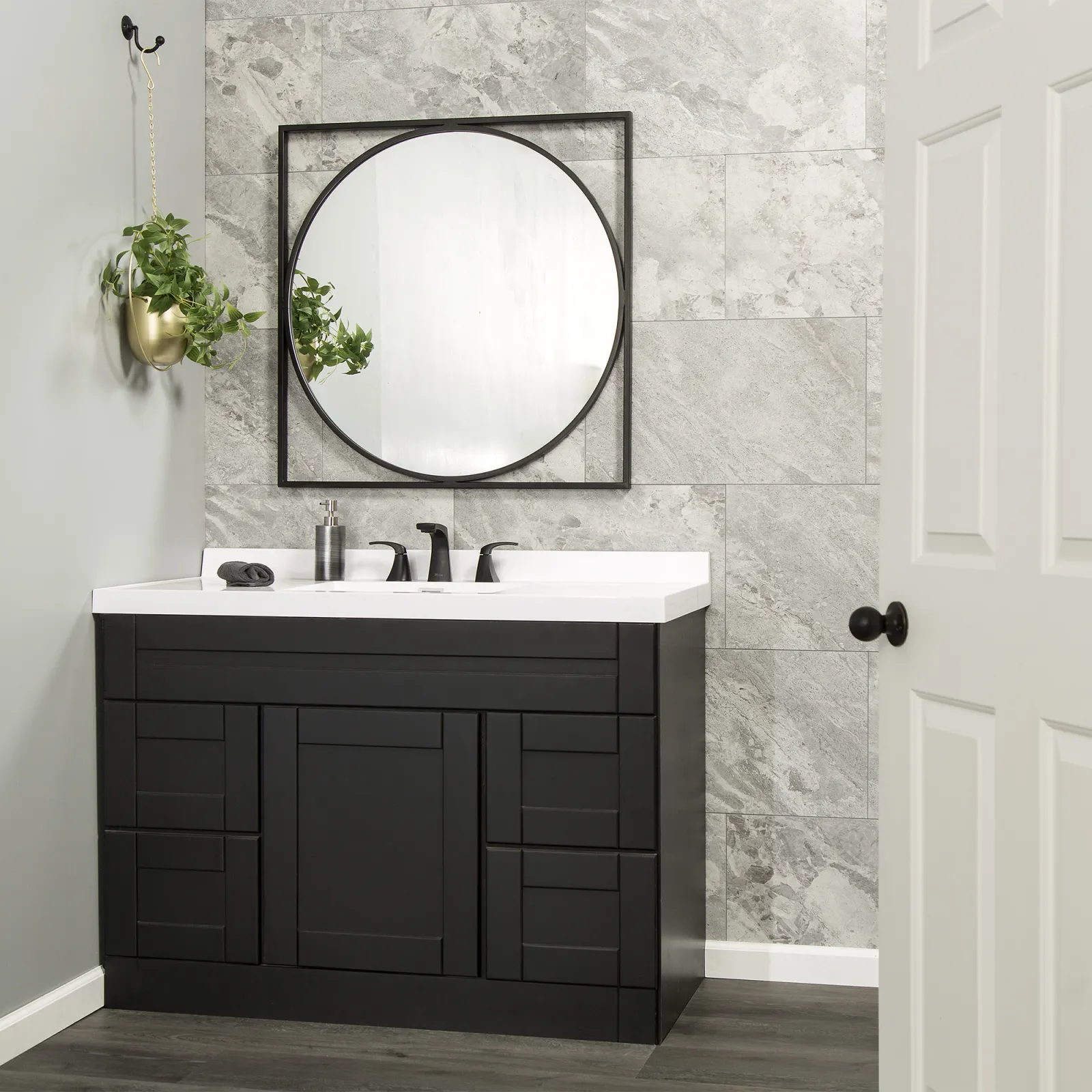 Modern black bathroom vanity with white countertop, round mirror, gray tiled wall, and hanging plant in gold pot.