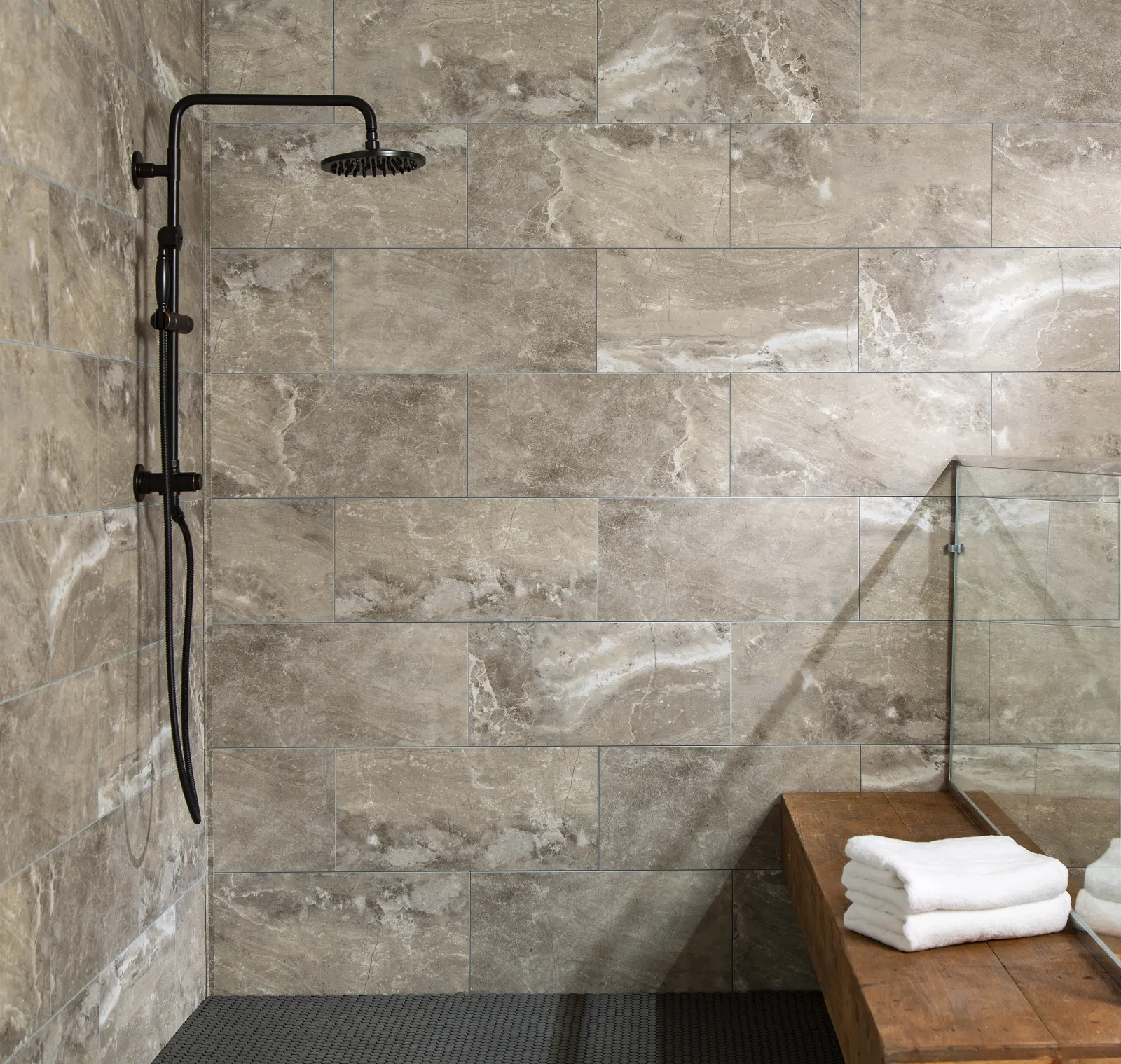Modern bathroom shower area with beige stone tiles, black rainfall showerhead, and wooden bench with white towels.