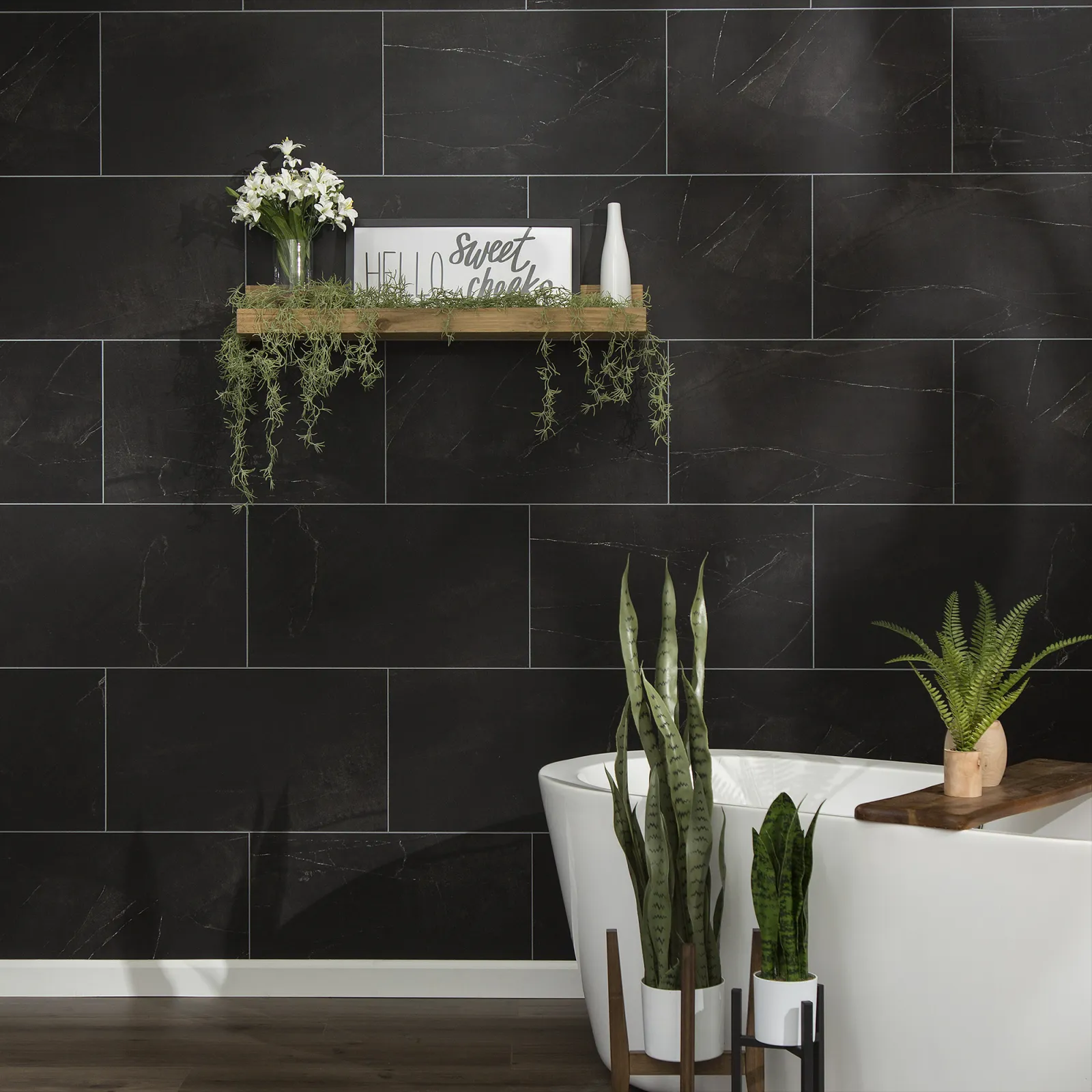 Modern bathroom with black marble tiles, white freestanding tub, wooden shelf with plants and decor.