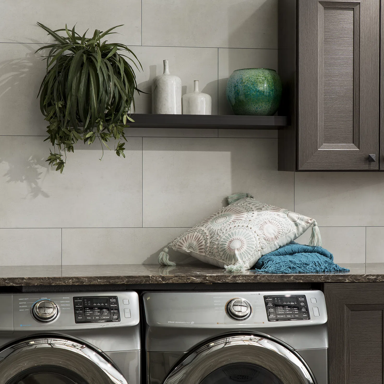 Modern laundry room with stainless steel washer and dryer, decorative pillows, hanging plant, and ceramic vases.