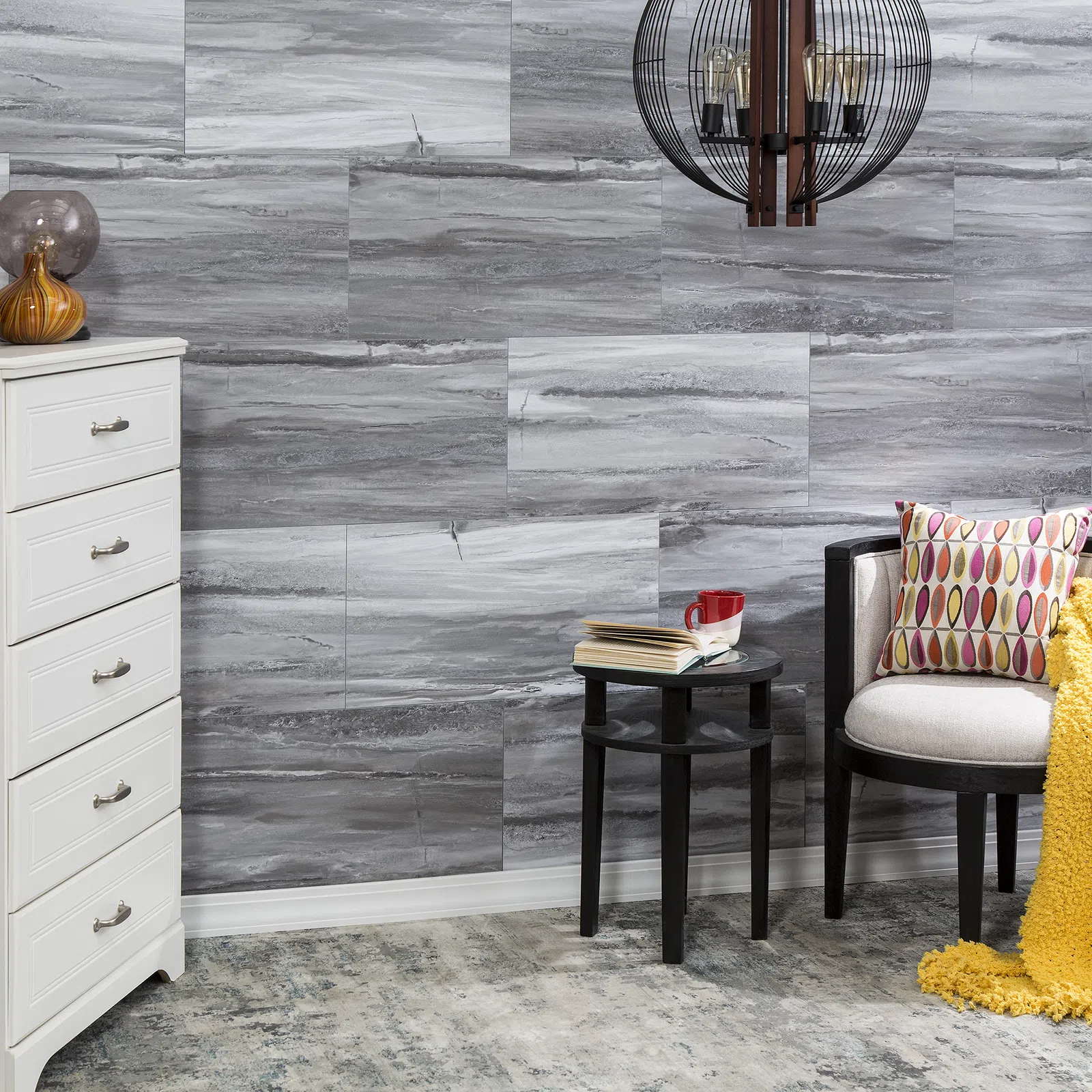 Cozy corner with white dresser, patterned pillow chair, black side table, gray stone wall, and decorative lighting fixture.