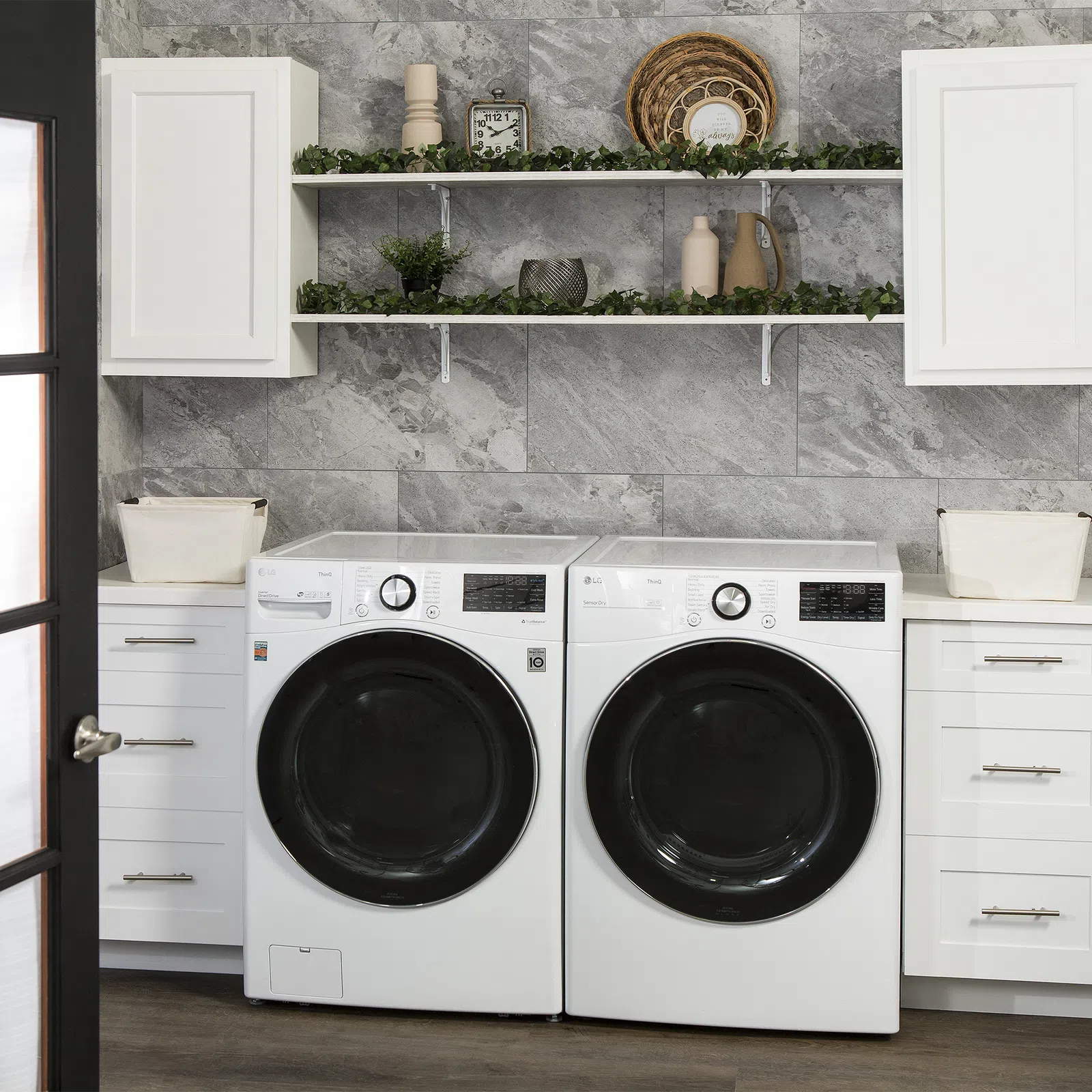 Modern laundry room with front-load washer and dryer, white cabinetry, grey stone wall, and decorative shelves.