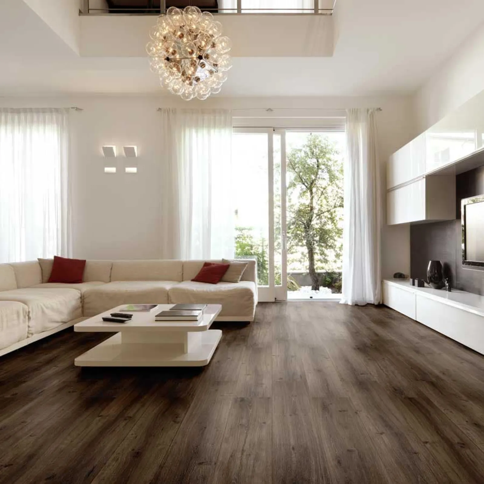 Modern living room with dark wood flooring, white sectional sofa, glass chandelier, and large windows with sheer curtains
