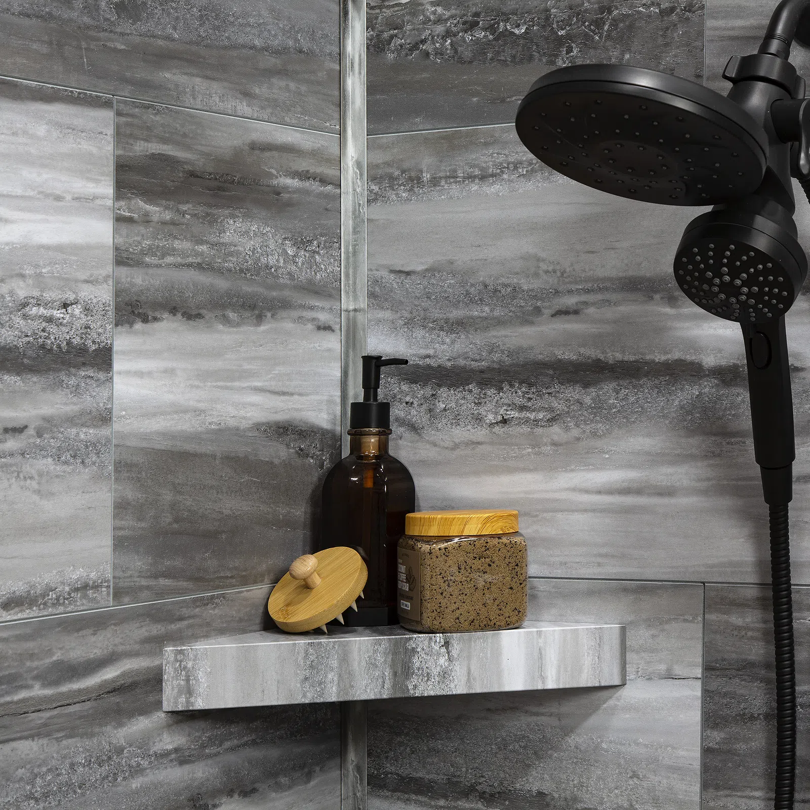 Modern bathroom shower with dark showerhead, gray marble tiles, soap dispenser, scrub brush, and exfoliating scrub jar.
