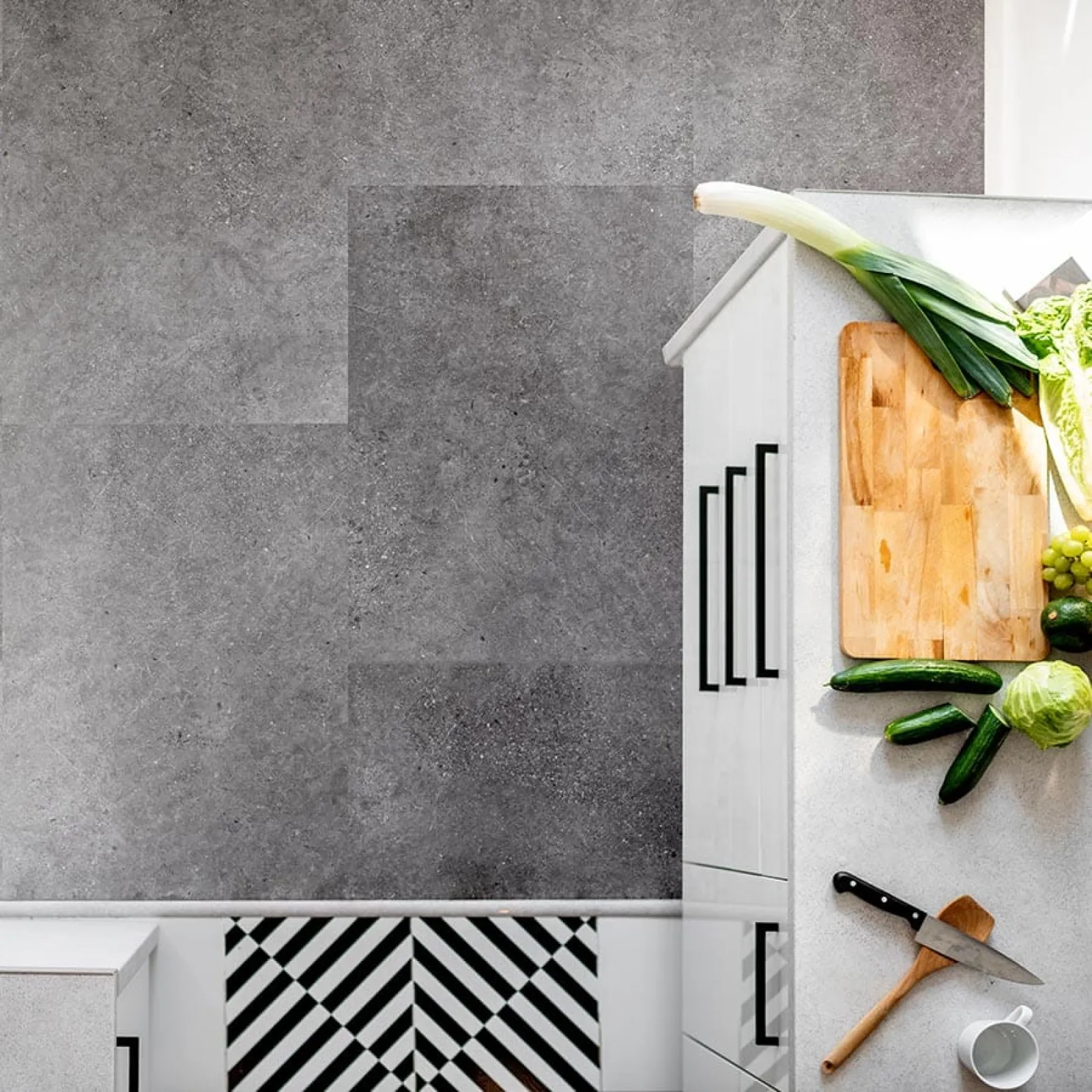 Top view of a white kitchen counter with chopping board, vegetables, knife, and modern gray tile flooring
