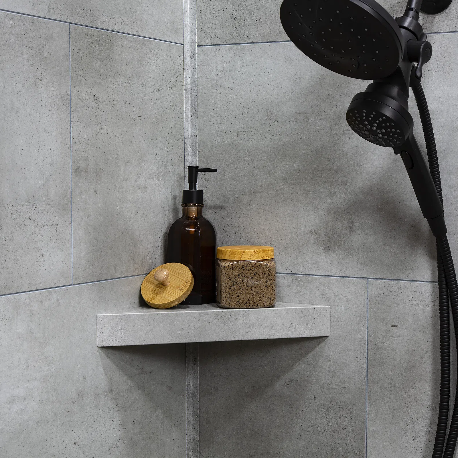 Modern shower corner with black showerhead and natural skincare containers on grey stone shelf.