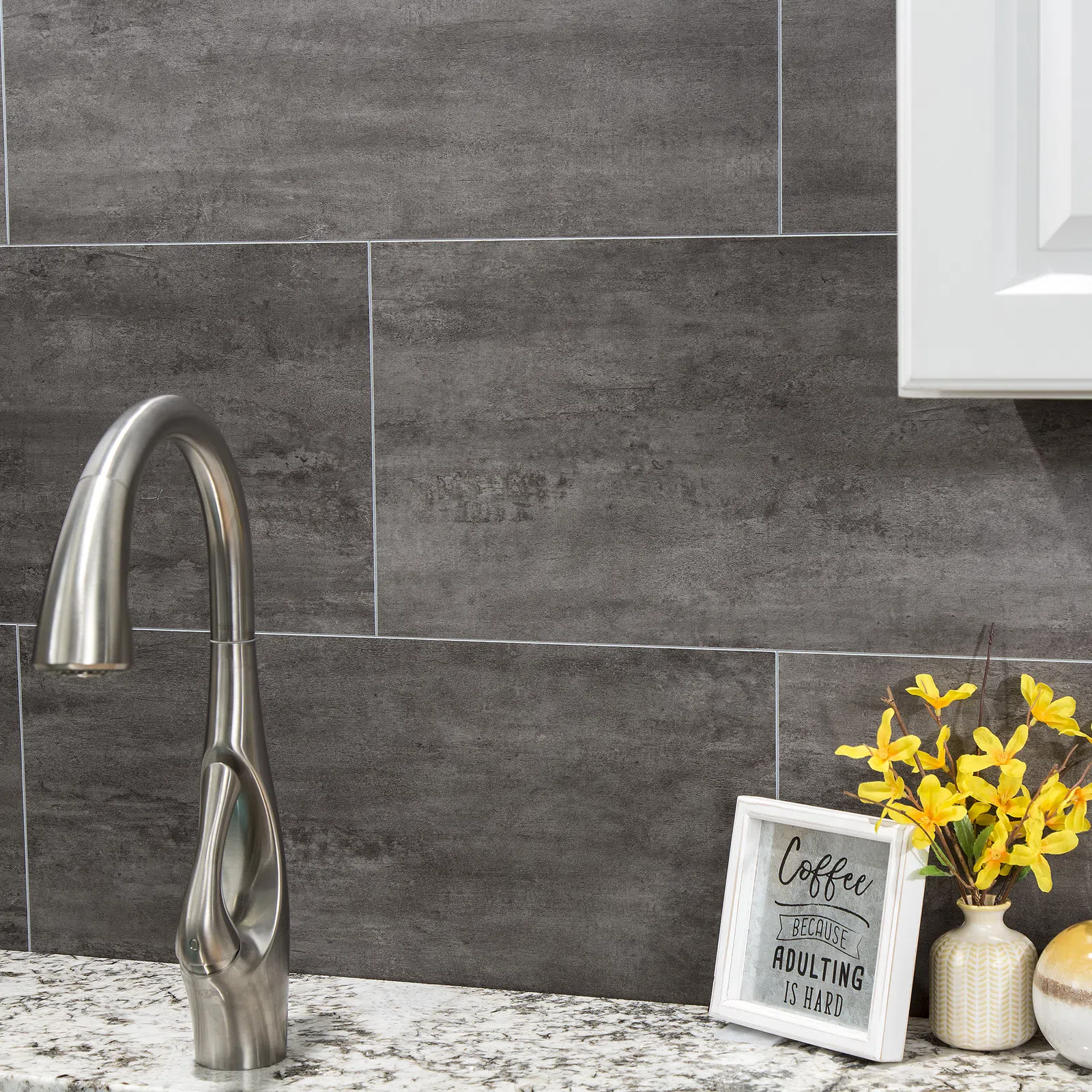 Modern kitchen faucet on granite countertop with dark tile backsplash and decorative items including framed coffee quote and yellow flowers.