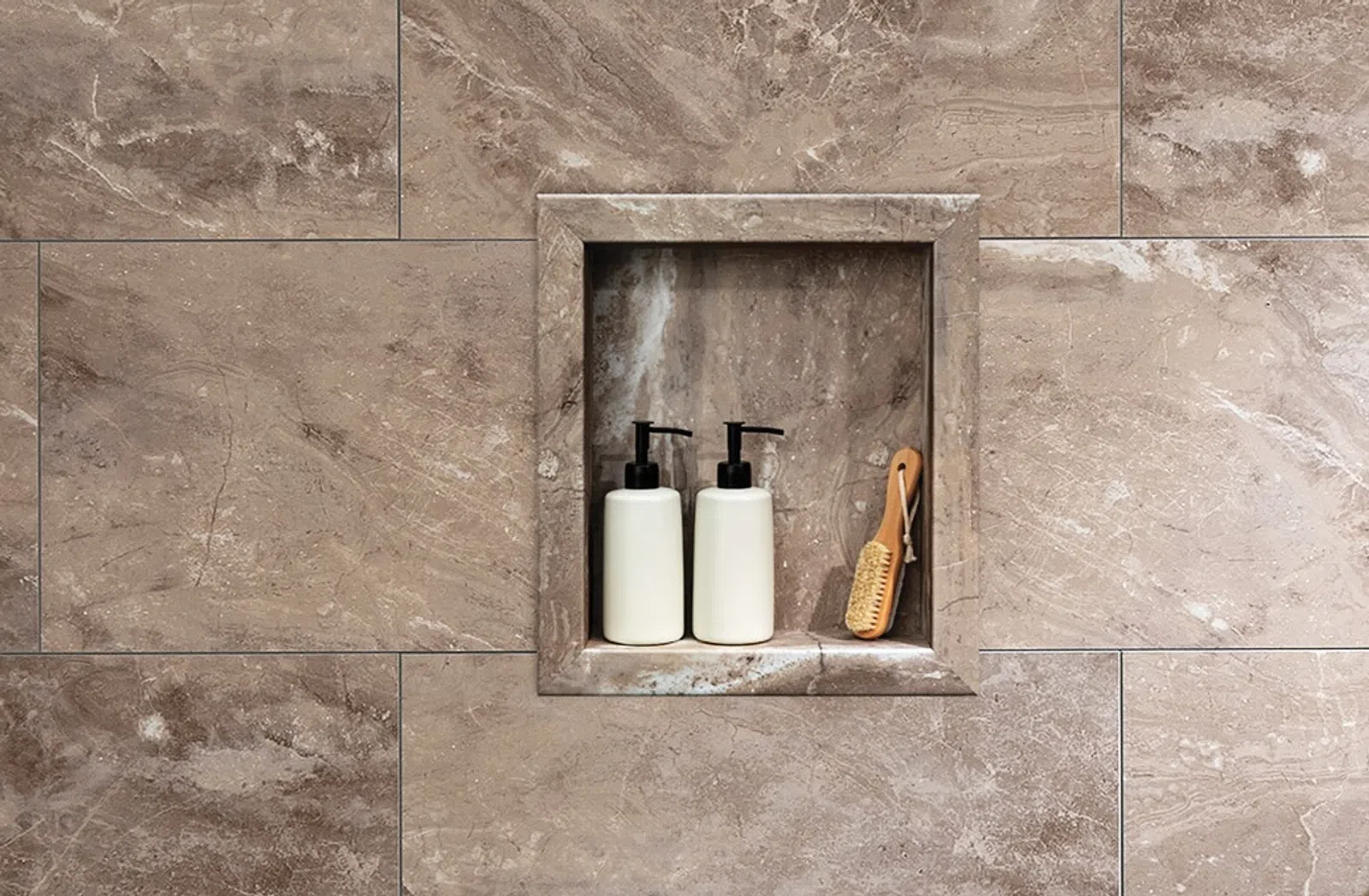 Two white pump bottles and a wooden brush placed in a stone wall niche with beige marble tiles around.