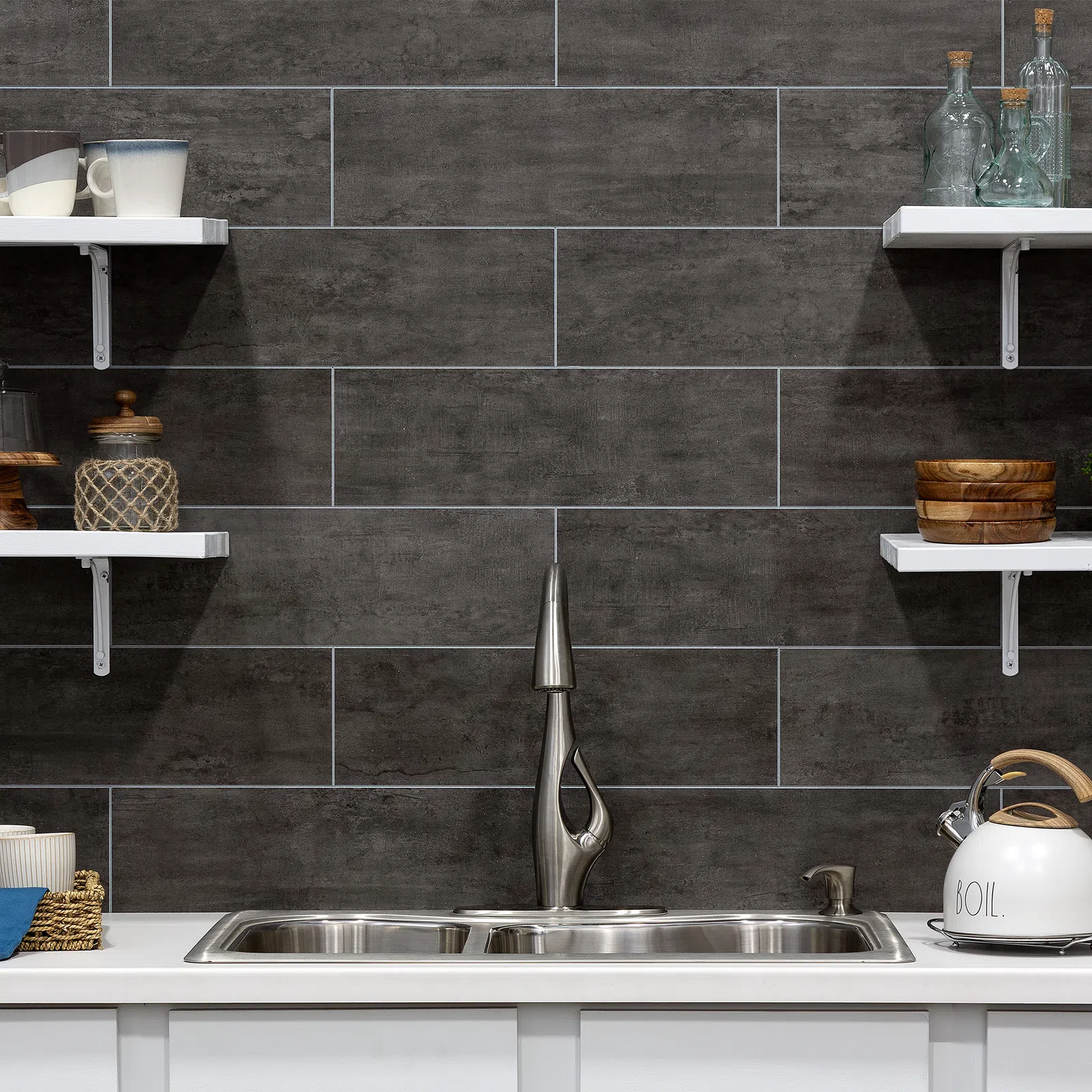 Modern kitchen sink with sleek faucet, dark textured tile backsplash, white shelves with mugs and glass bottles.