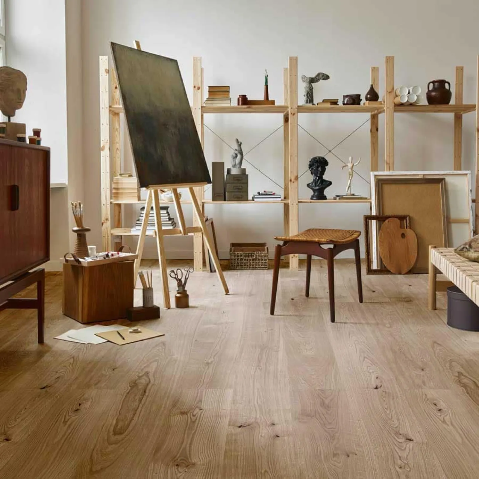Cozy art studio with wooden flooring, easel, sculptures, wooden chair, and open shelves filled with art supplies.