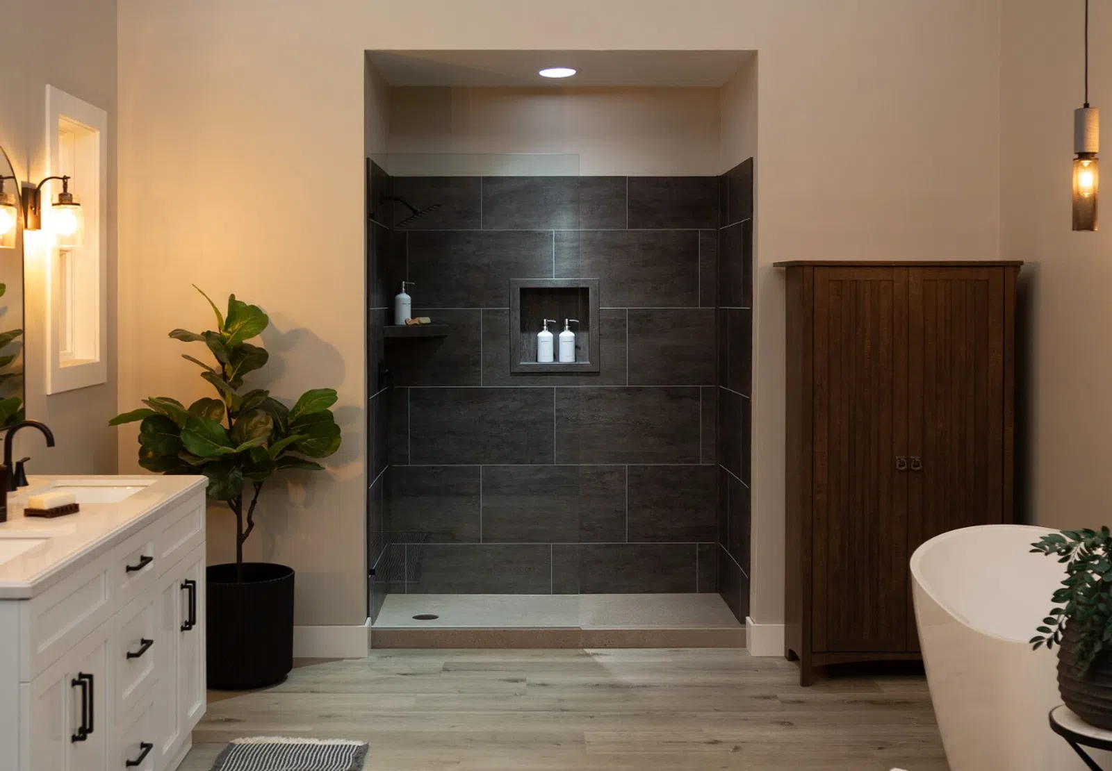 Modern bathroom with white vanity, freestanding tub, dark tile shower, wooden cabinet, and ambient lighting.