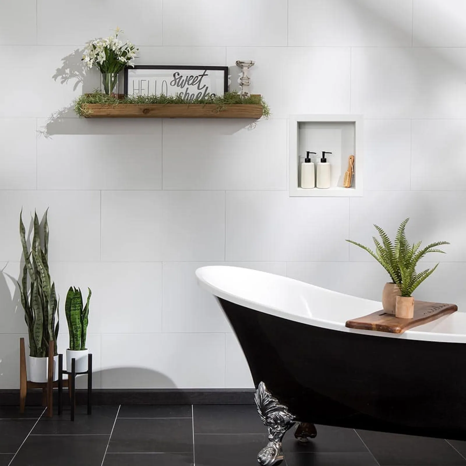 Modern bathroom with black clawfoot bathtub, white tiled walls, plants, and decorative shelf with framed sign.