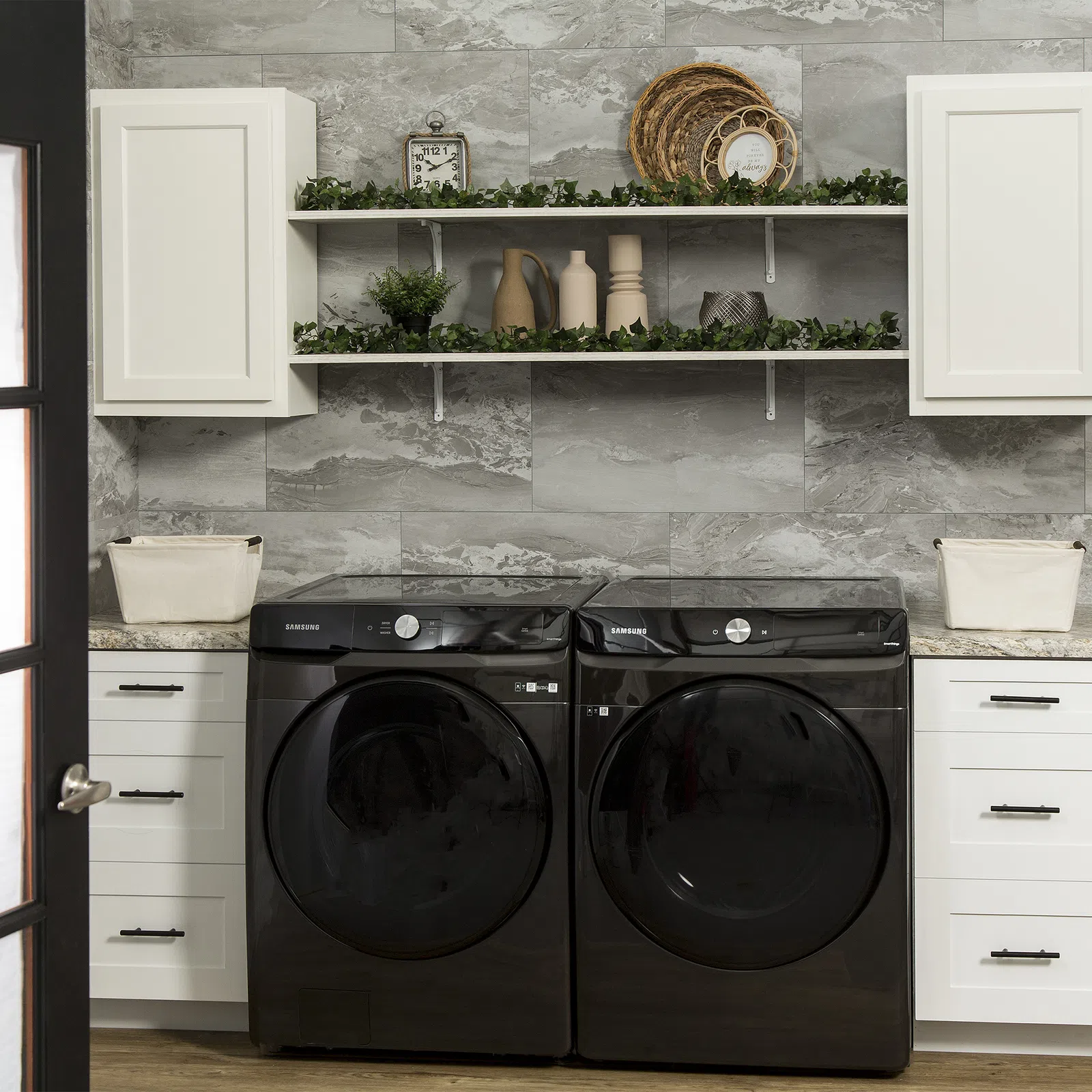 Modern laundry room with black Samsung washer and dryer, white cabinets, decorative shelves, and gray tile wall.