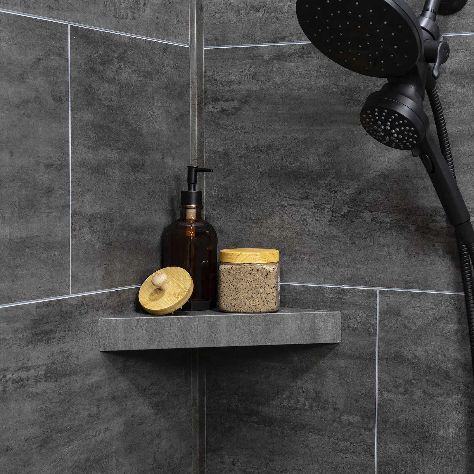 Modern shower corner with dark gray tiles, black showerhead, amber soap dispenser, and scrub jar with wooden lids.