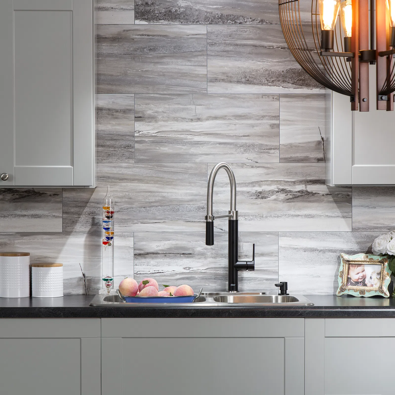 Modern kitchen sink with black faucet, marble backsplash, peaches in blue tray, and decorative jars on countertop.