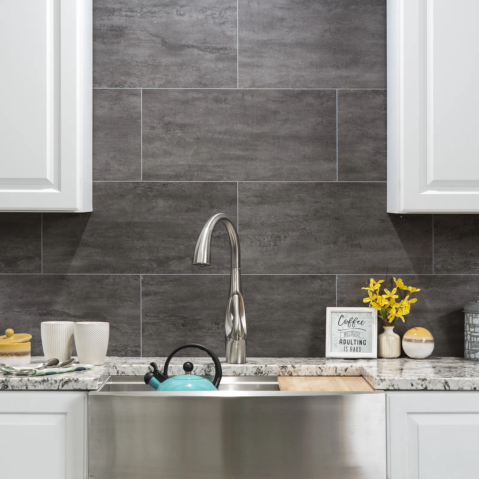 Modern kitchen sink with stainless steel faucet, turquoise kettle, grey tile backsplash, and yellow flowers.