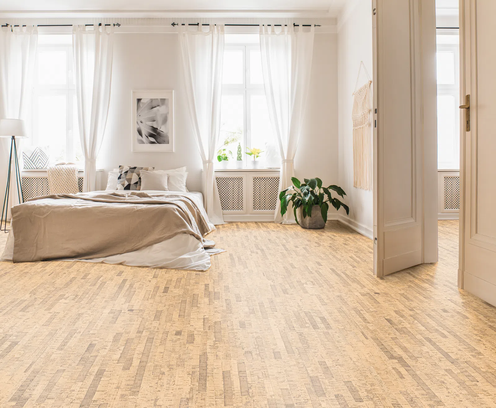 Bright minimalist bedroom with large windows, white curtains, beige bedding, wooden floor, and indoor plant by doorway.