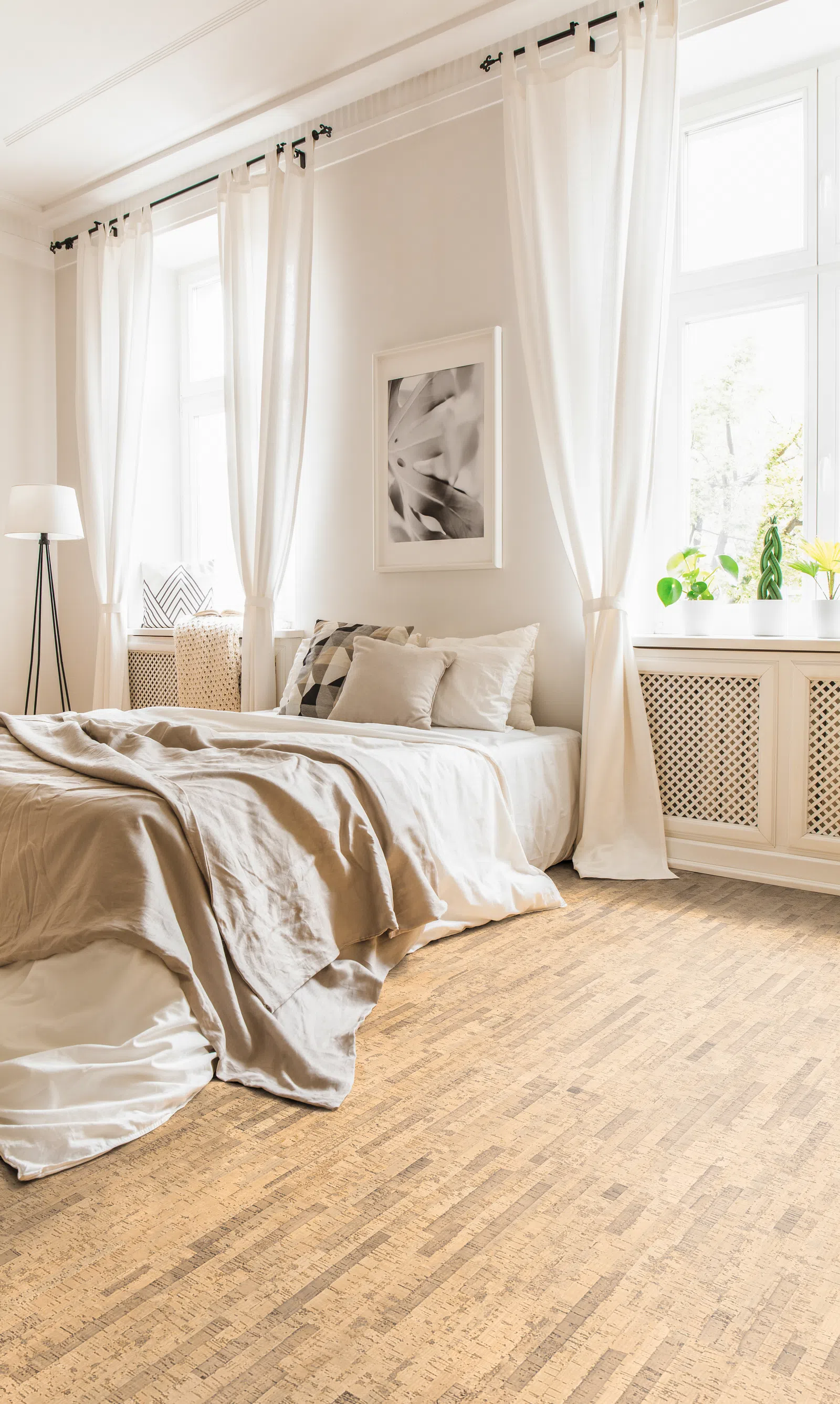 Bright modern bedroom with beige linen bedspread, white curtains, potted plants, and cork flooring in natural light.