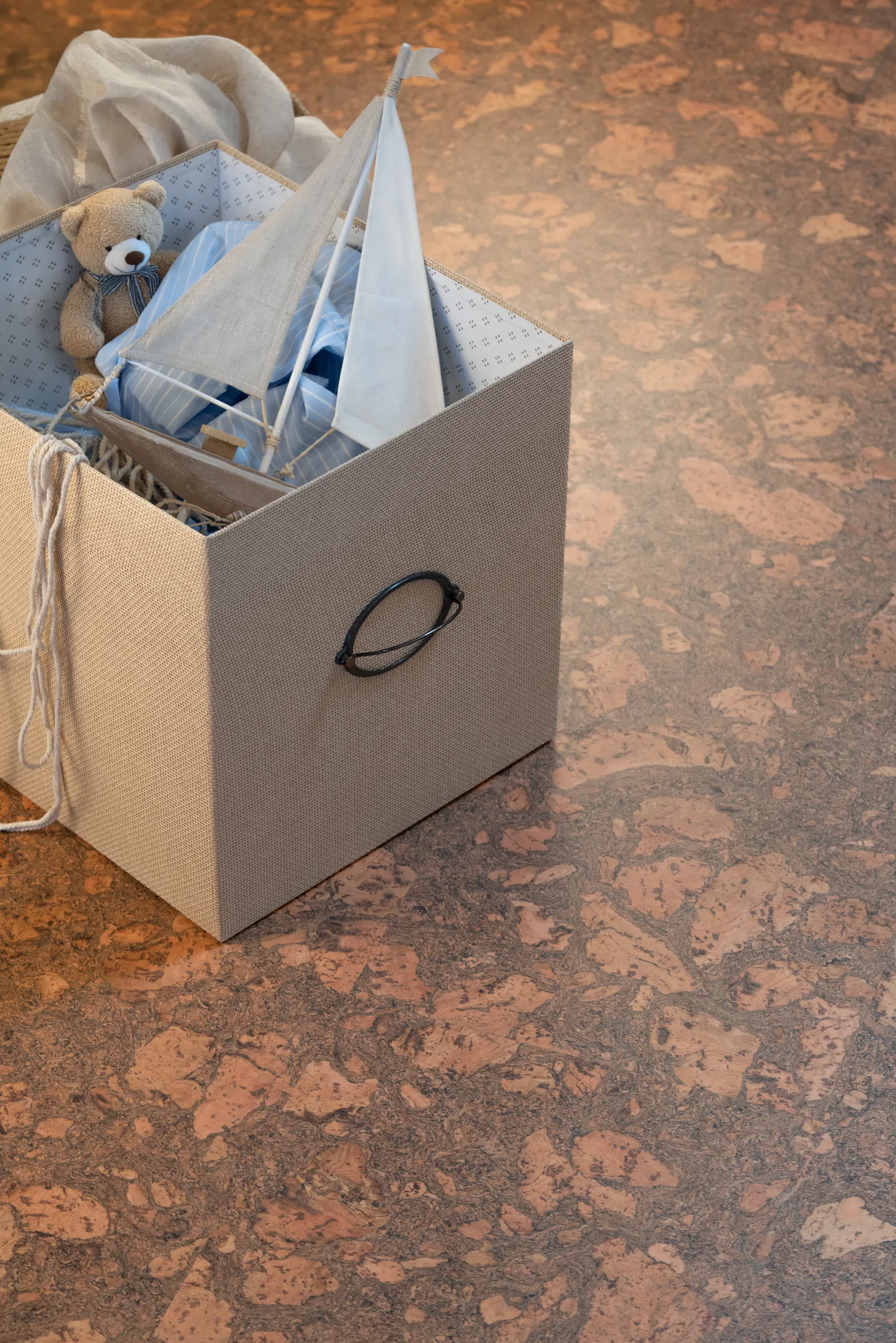 Beige storage box holding a teddy bear, fabric sailboat, and cloth on textured brown floor.