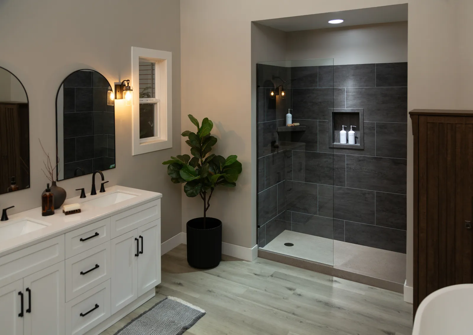 Modern bathroom with white double vanity, black fixtures, indoor plant, and walk-in shower with dark tiles and glass door