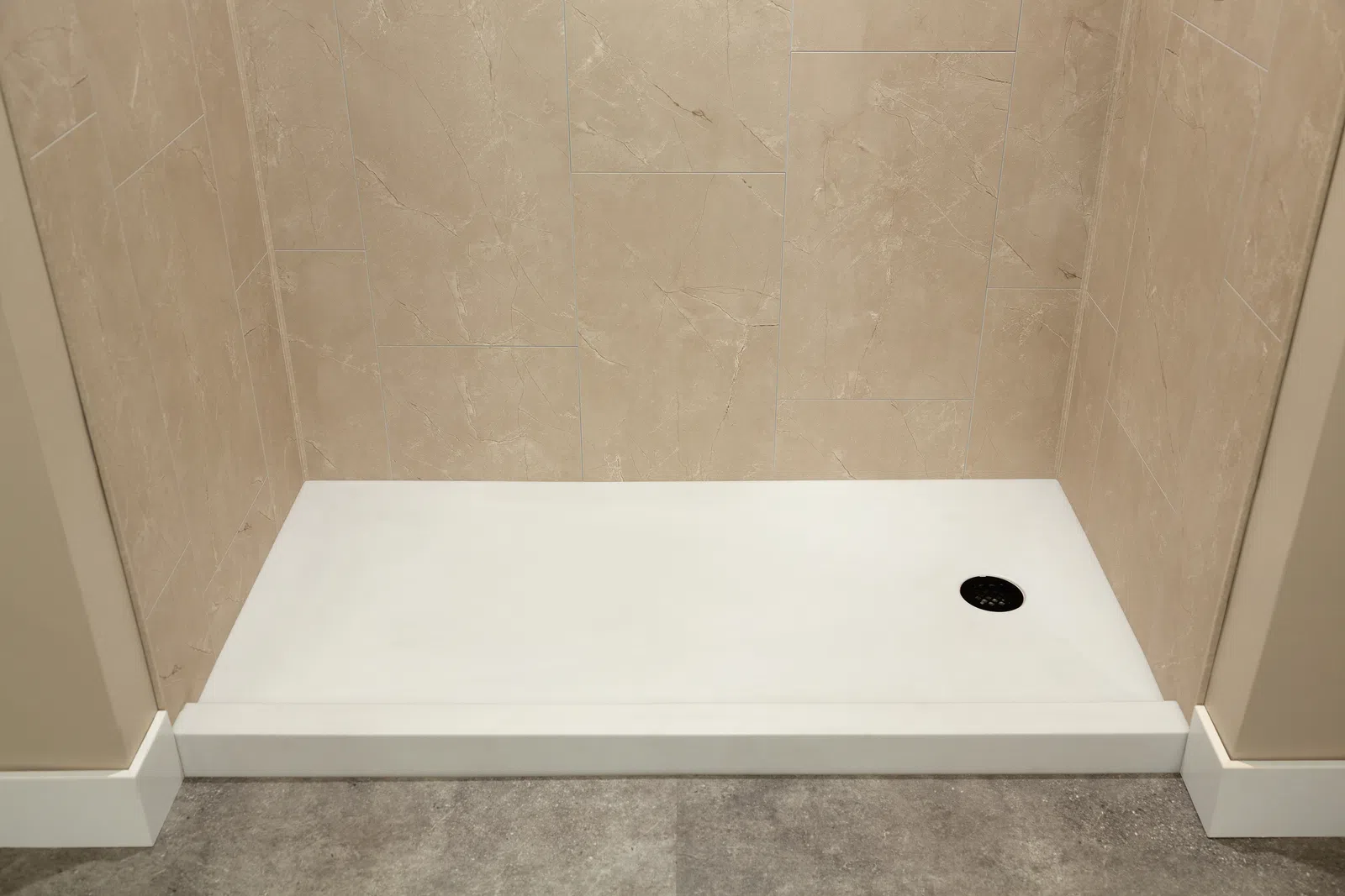 Minimalist shower base with beige tiled walls and gray stone floor featuring a circular black drain.